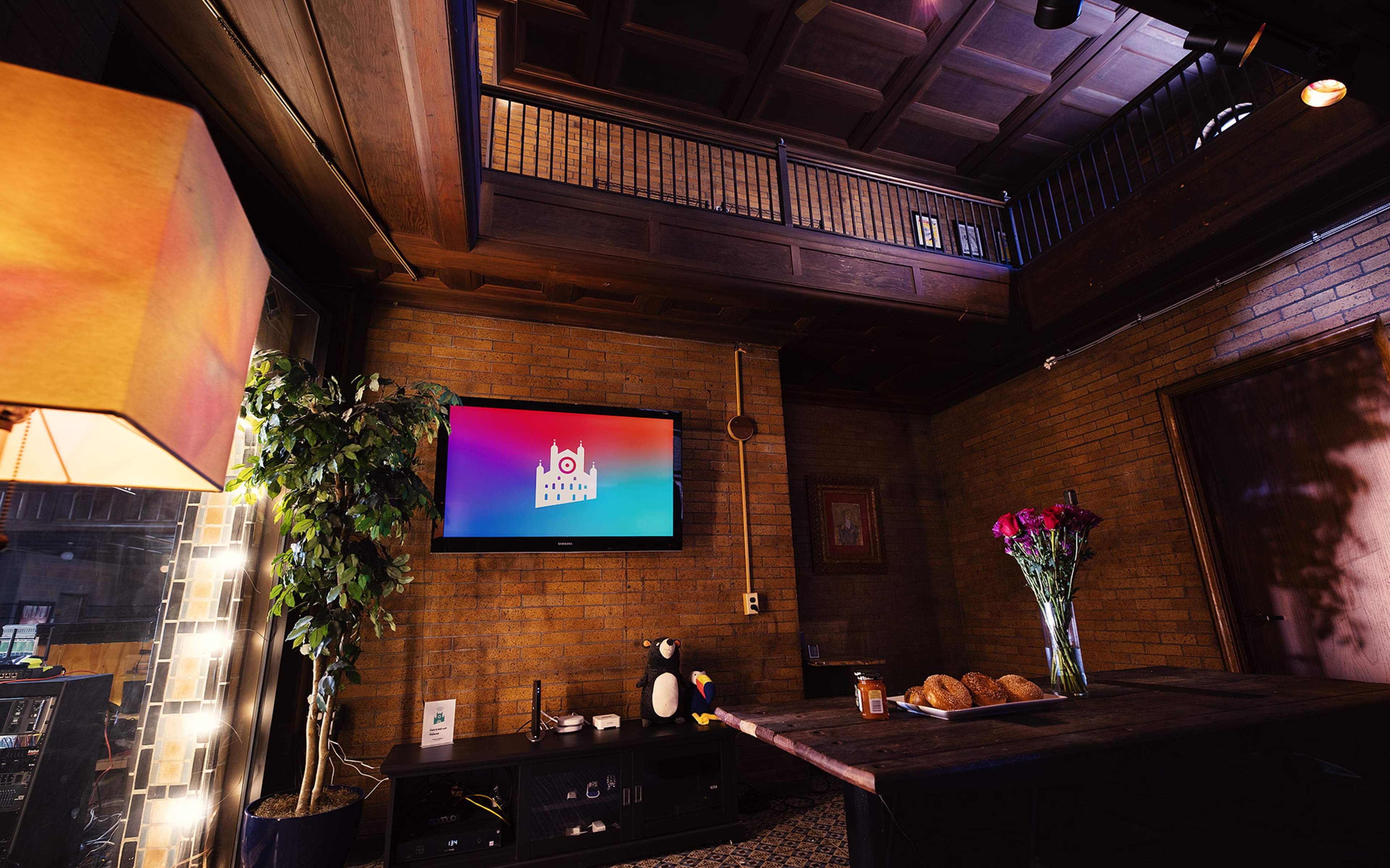 A cozy room with a brick wall, a wooden ceiling, a television displaying a logo, a table with croissants and flowers, and a plant in the corner.