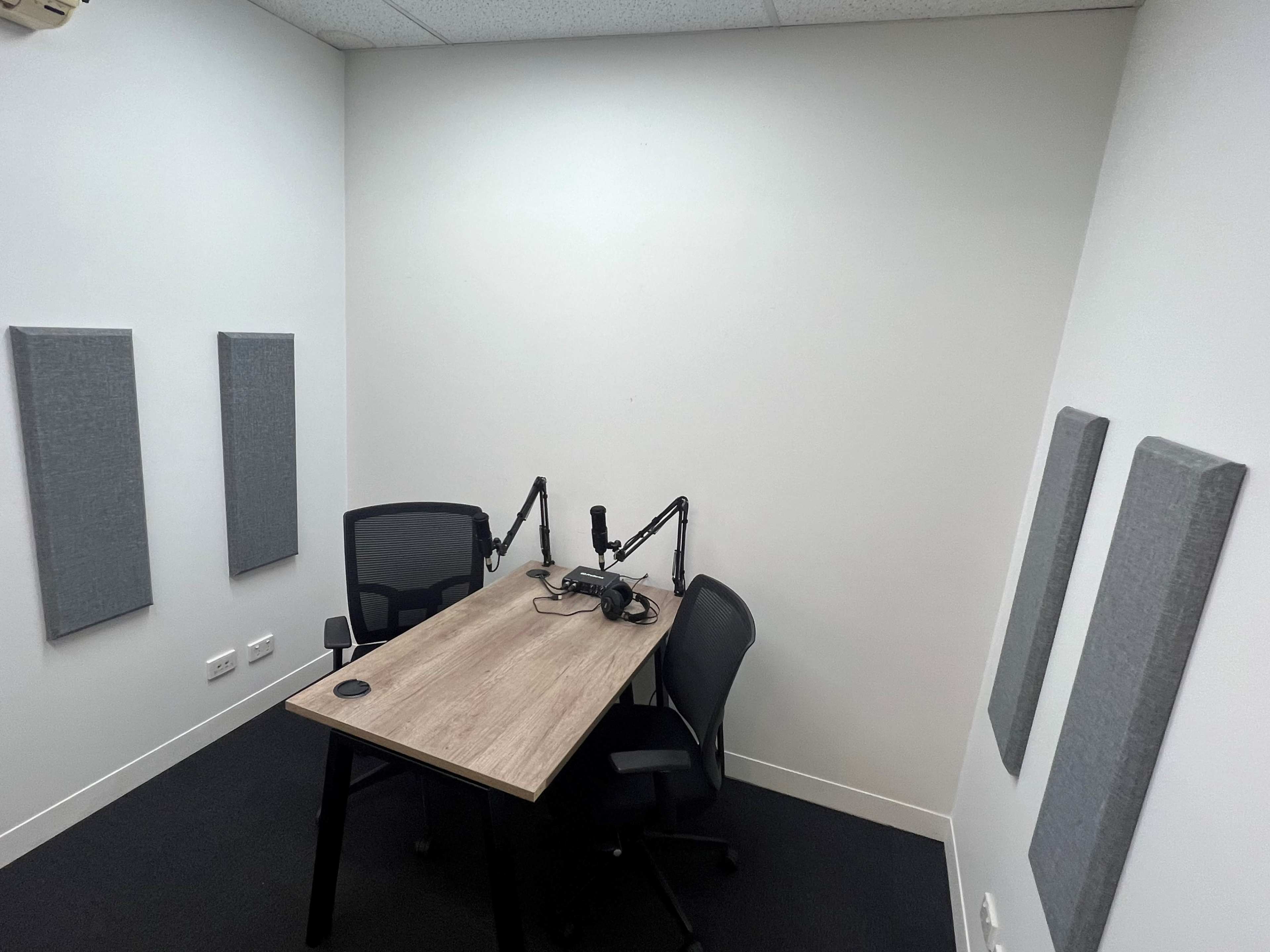The image shows a small, empty recording studio with a wooden table, two chairs, and microphones mounted on adjustable arms.