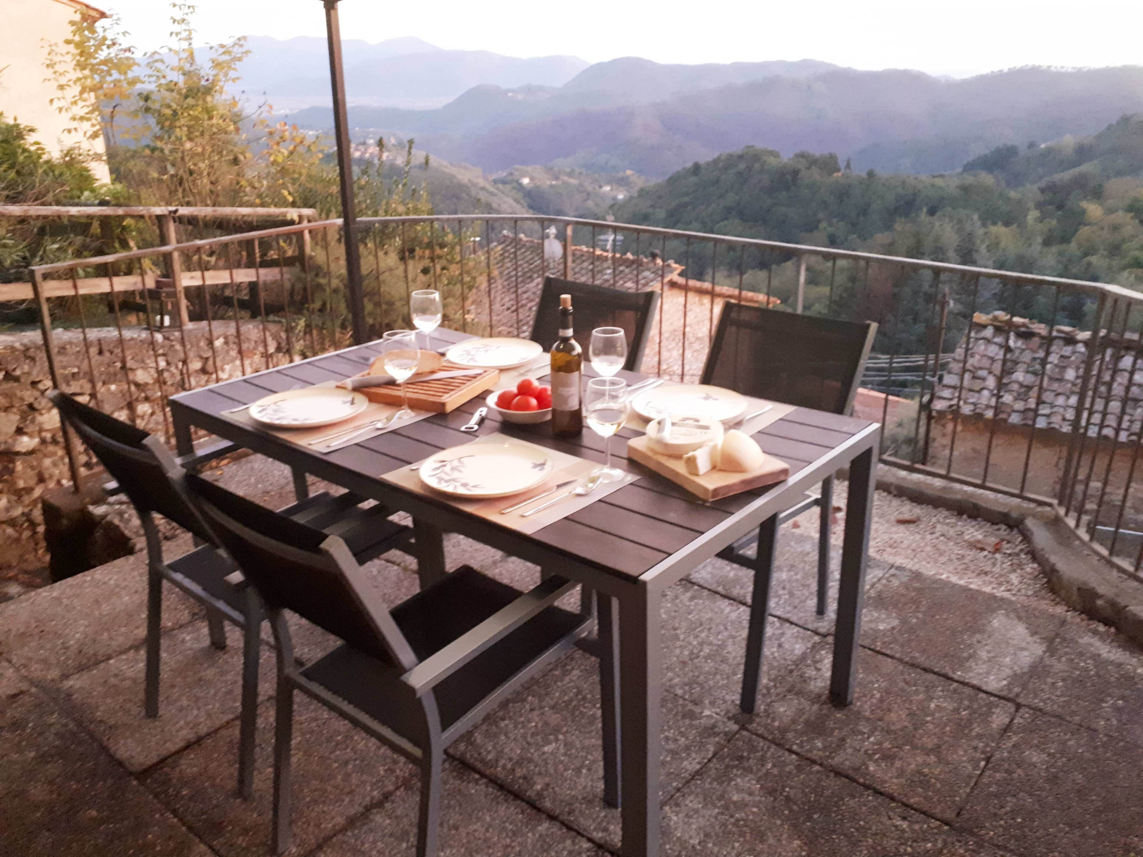 A dining table set with plates, utensils, glasses, and food overlooks a mountainous landscape during twilight.
