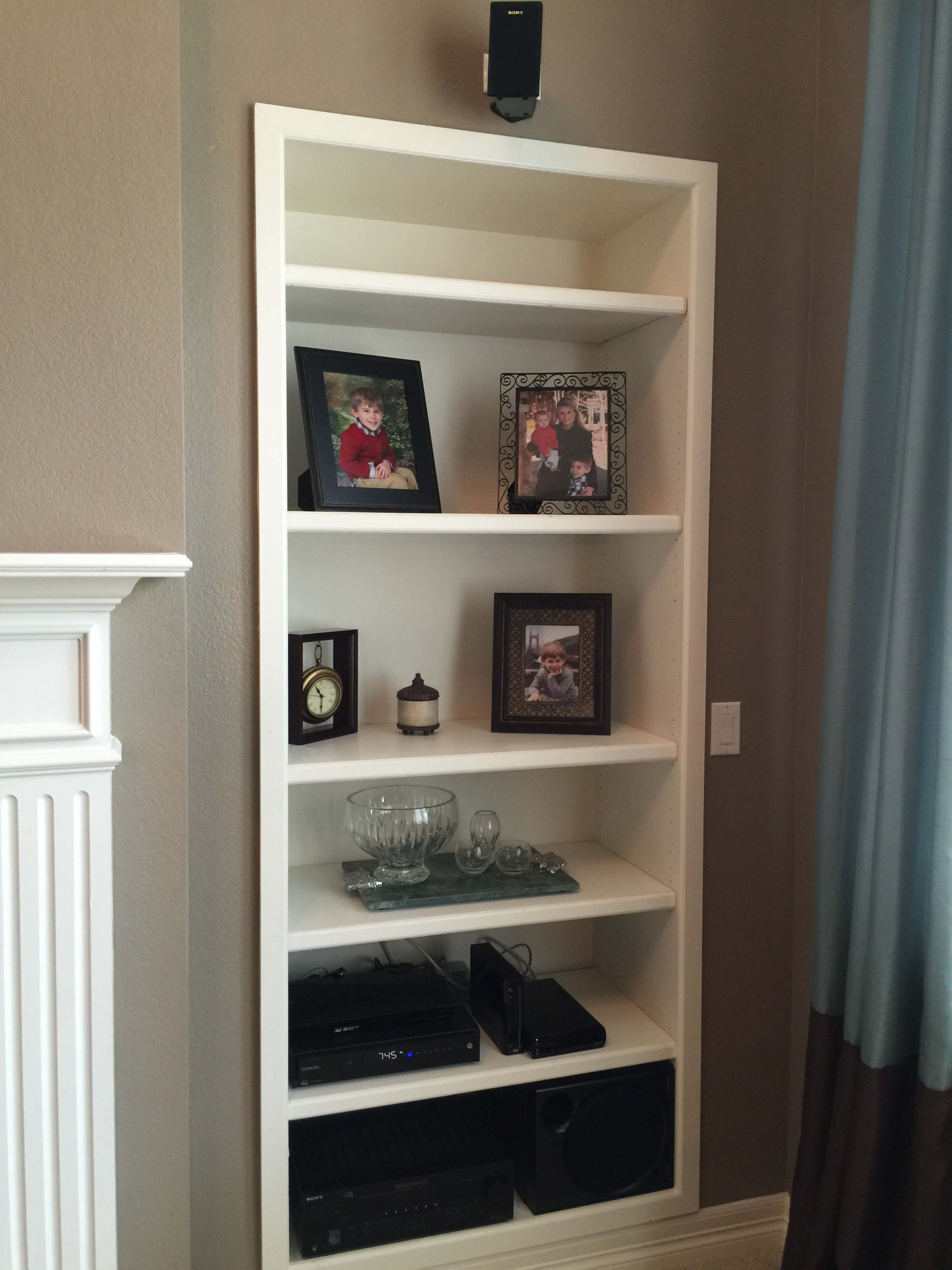 A white bookshelf with several shelves displaying framed photographs, a clock, a glass dish, and electronics.