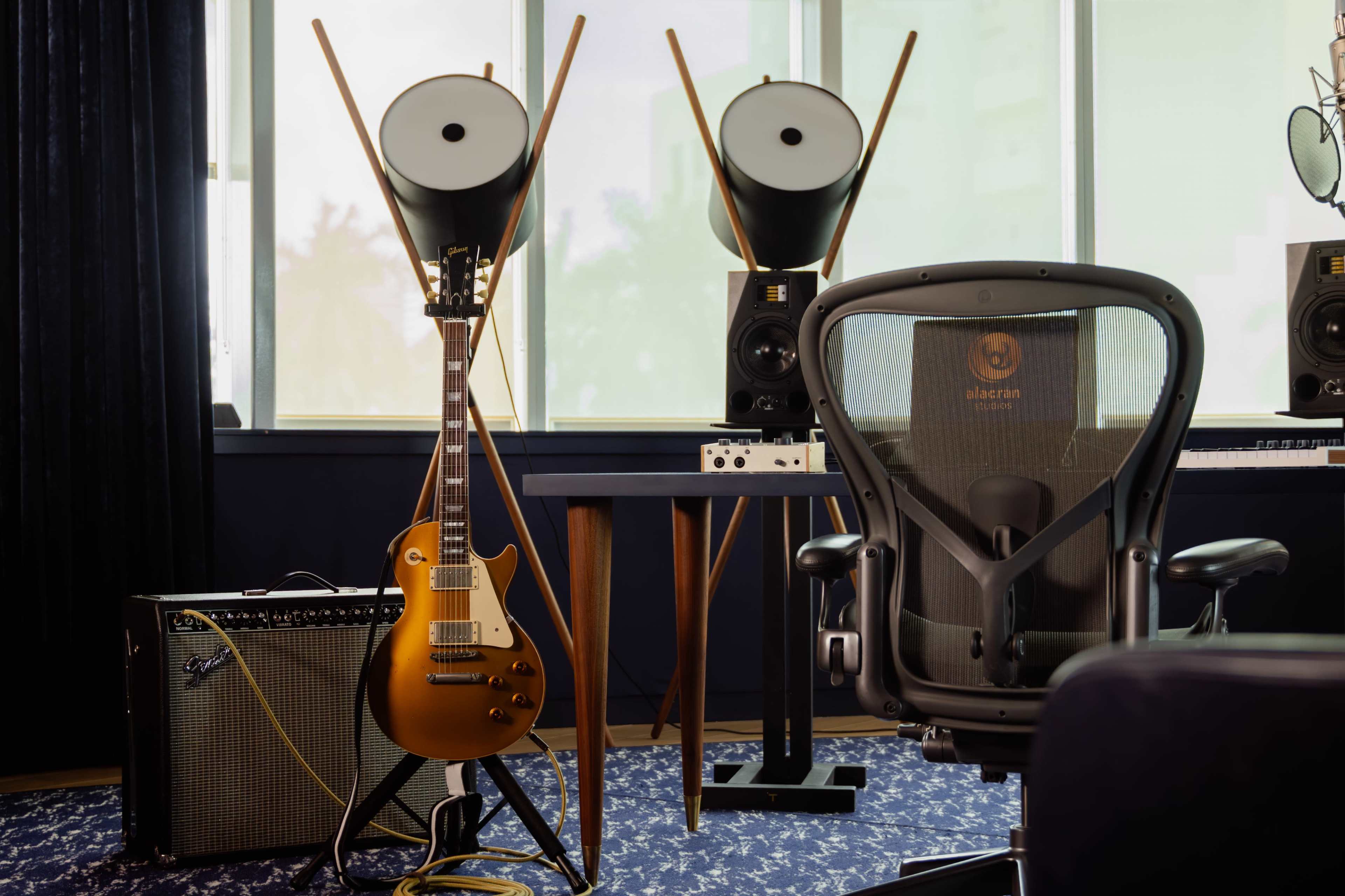 The image depicts a guitar resting against an amplifier next to a stylish office chair, with two speaker stands positioned in front of a large window.