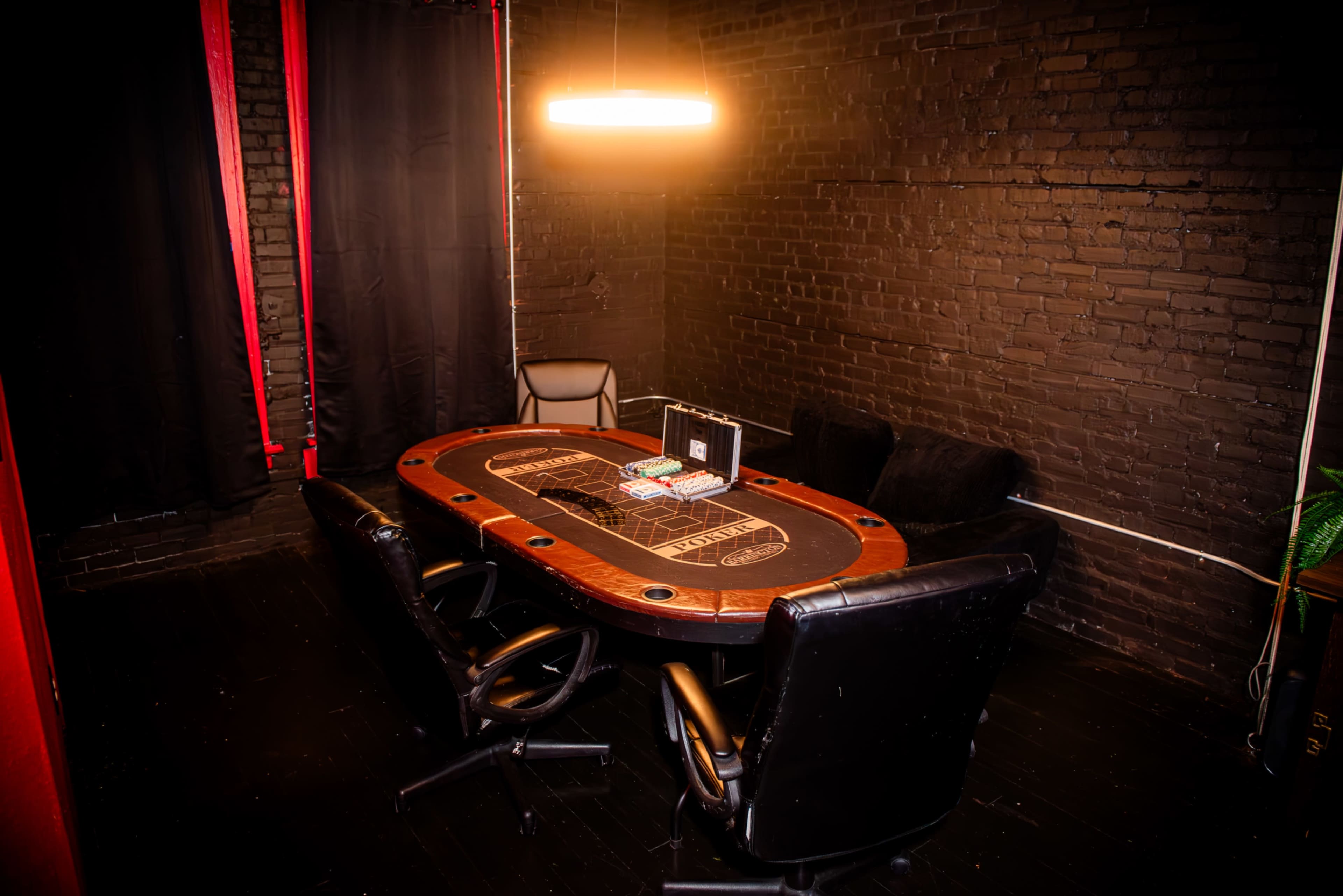 A poker table with four black chairs and a dealer's box is set up in a dimly lit room with exposed brick walls and red accents.