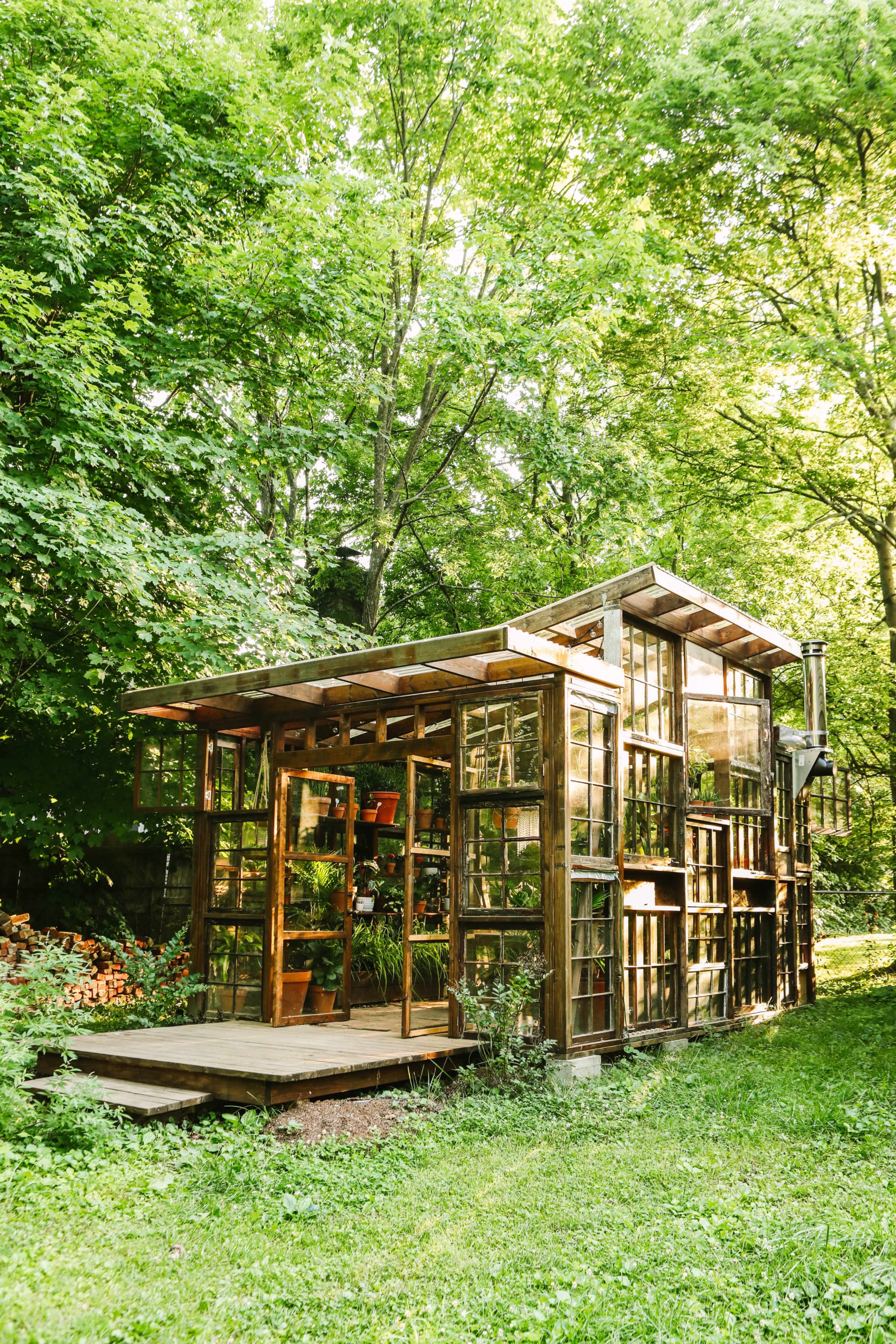 A wooden structure with numerous glass panels sits in a grassy area surrounded by trees.