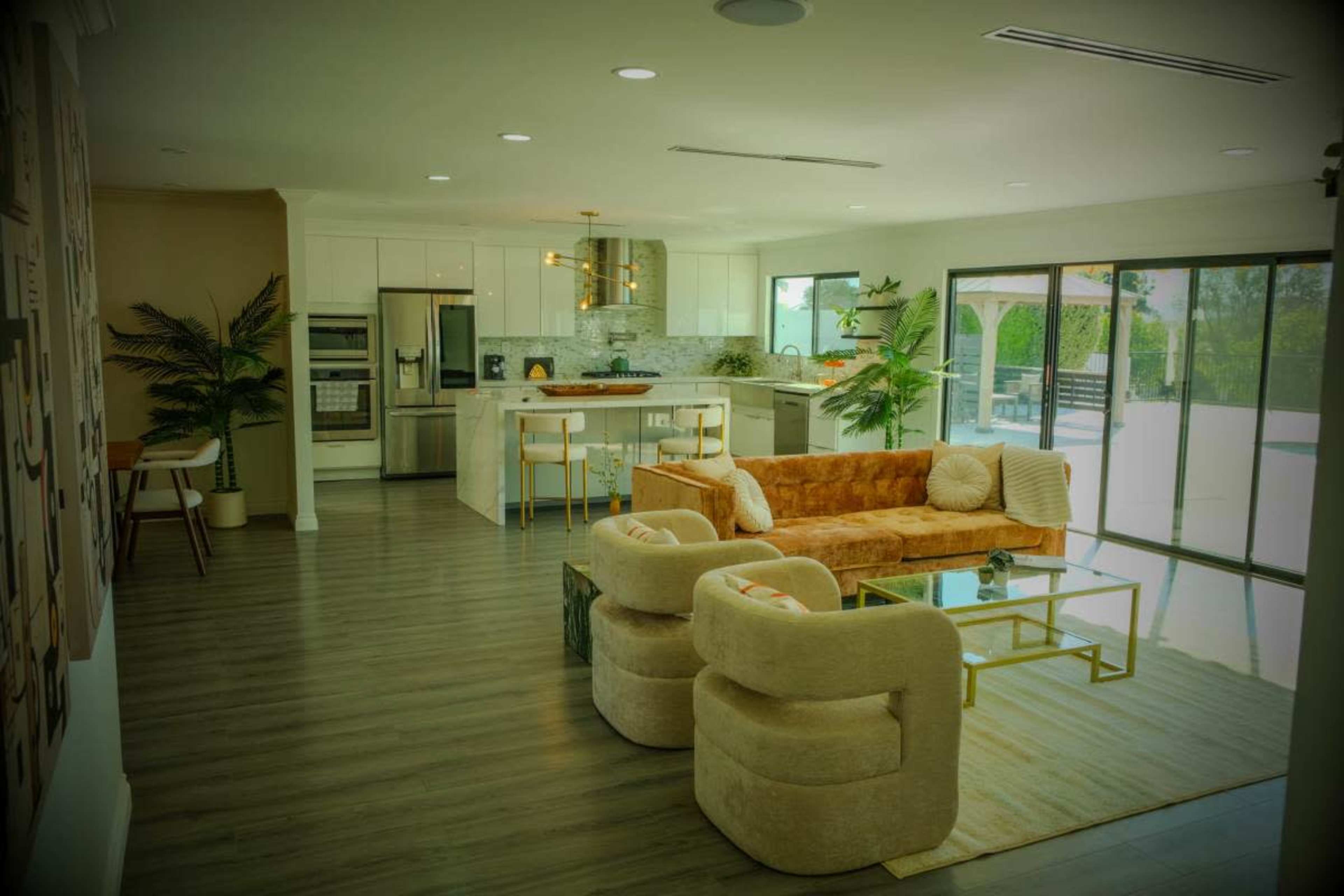 The image shows a modern open-concept living area featuring a cozy orange sofa and two plush chairs, with a kitchen and large glass doors leading to a patio in the background.