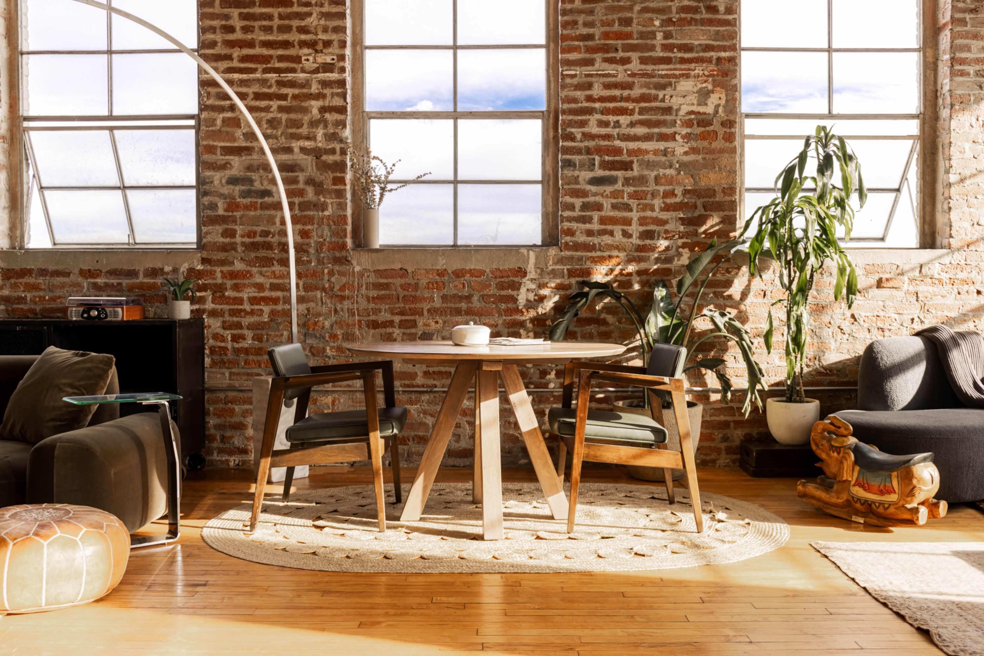 A rustic room with a round wooden table, two chairs, potted plants, and large windows allowing natural light to illuminate the space.