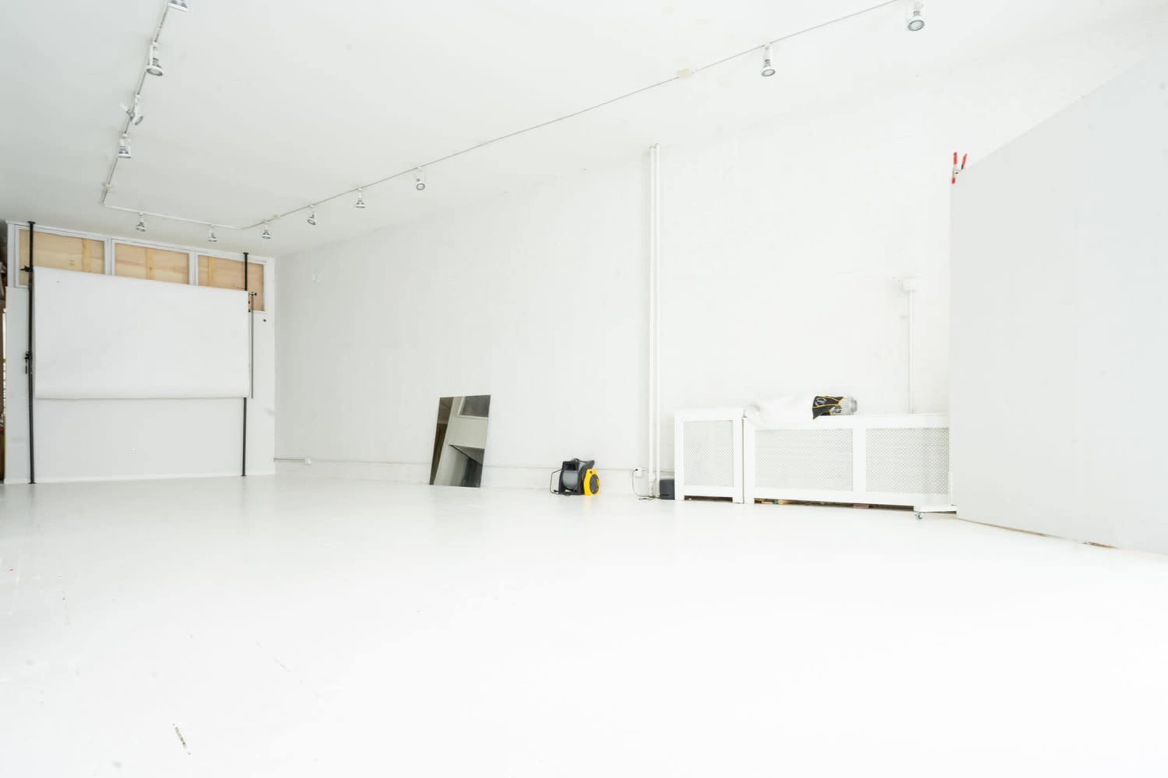 A spacious, empty room with white walls, a wooden platform, and minimal furniture, including a mirror and a fan.