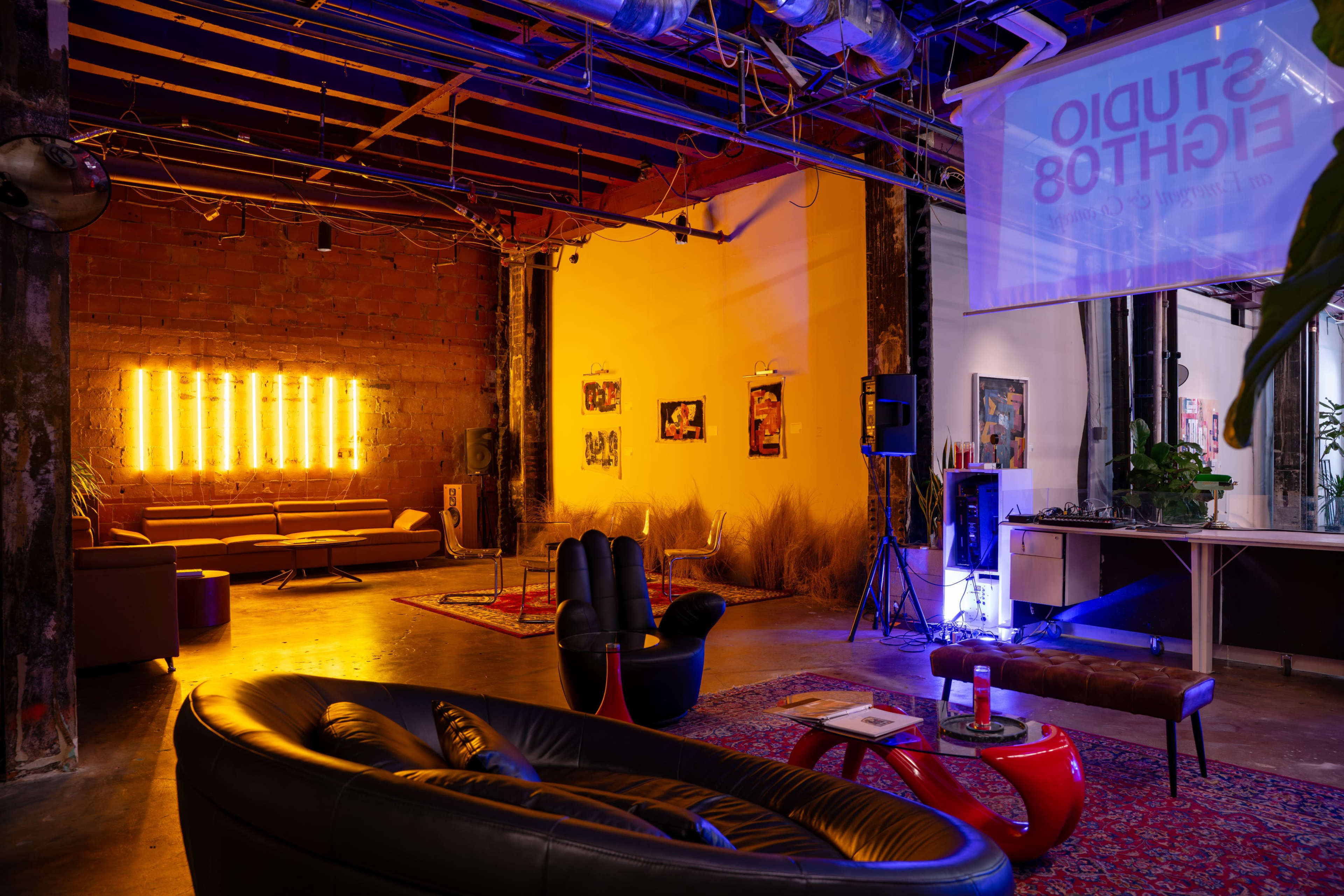 The image shows a modern lounge area featuring a mix of furniture, warm lighting, and artwork on the walls, set in an industrial space with exposed beams.