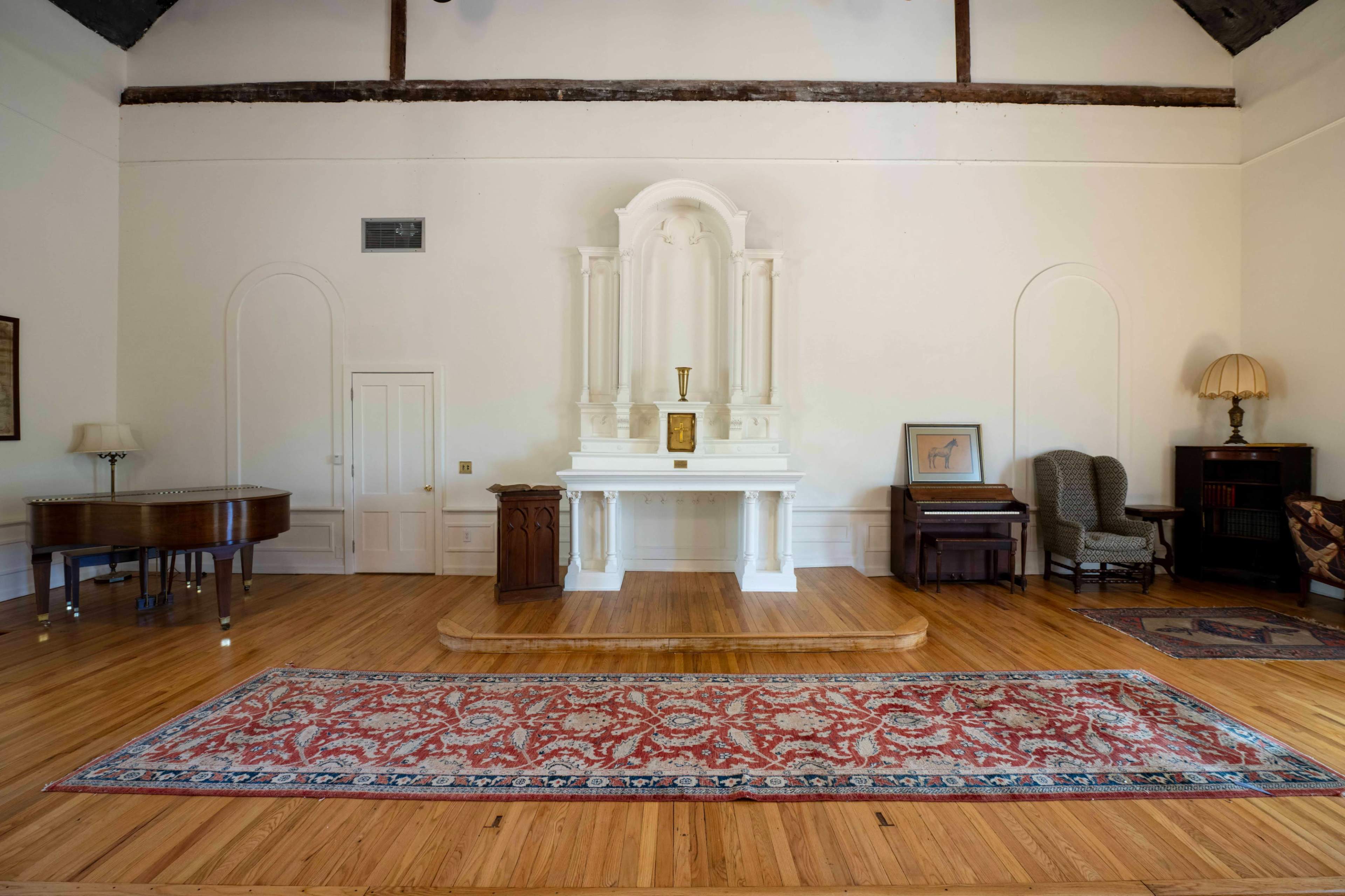 The image shows a spacious room with wooden flooring, featuring a white altar at one end, a grand piano, and various pieces of furniture, including a lamp and a bookshelf.