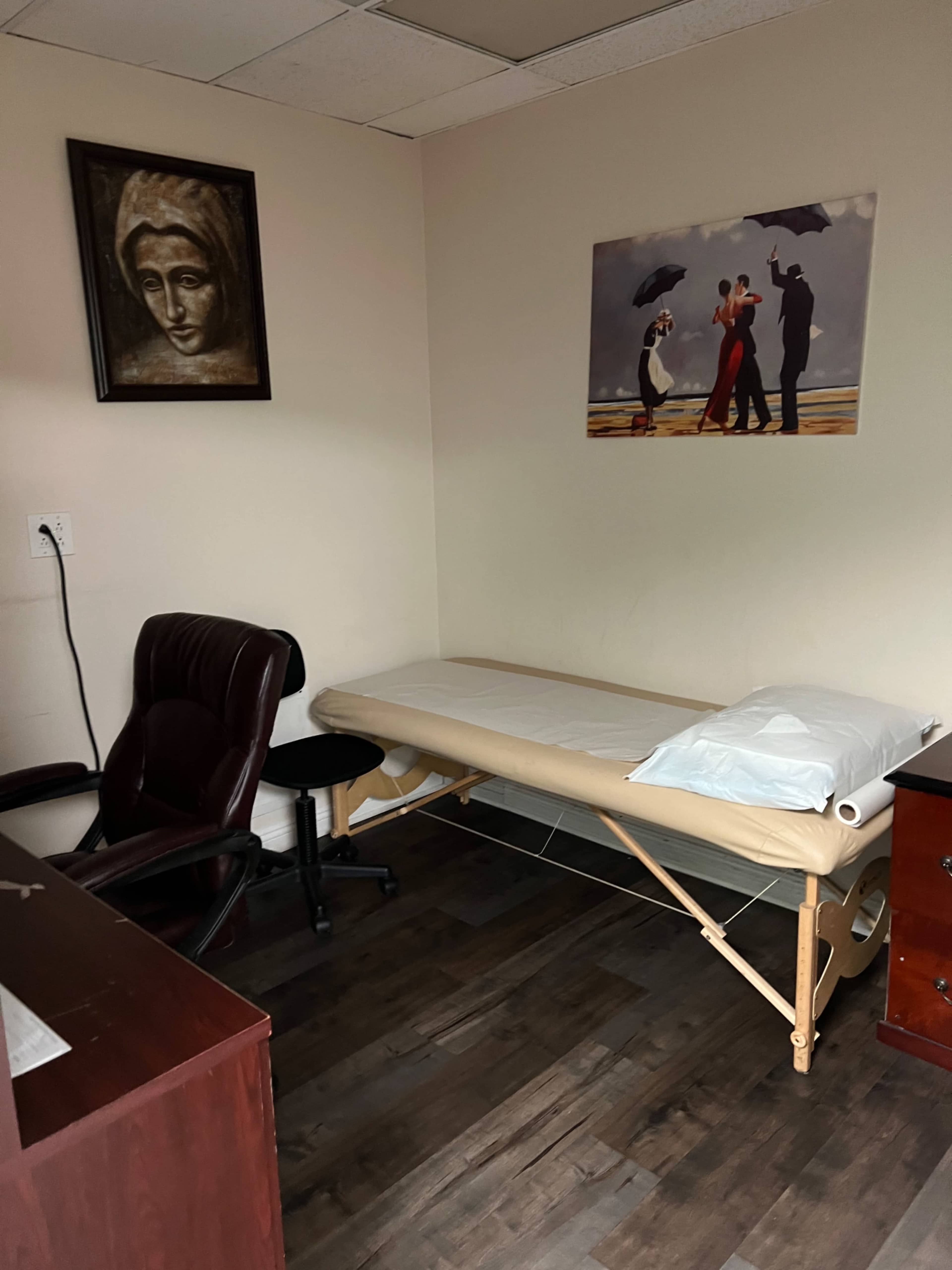 A small examination room with a massage table, a chair, a desk, and framed artwork on the walls.