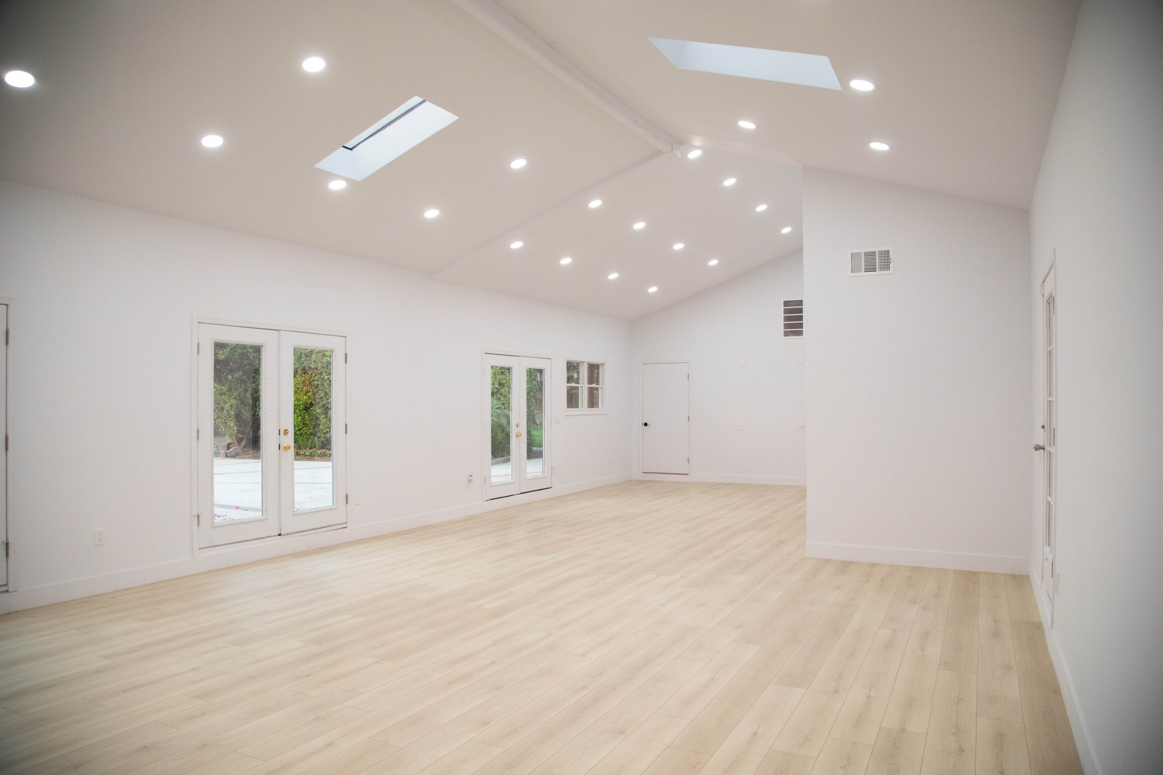 A spacious, empty room with a high ceiling, featuring large windows and sliding glass doors leading to an outdoor area.