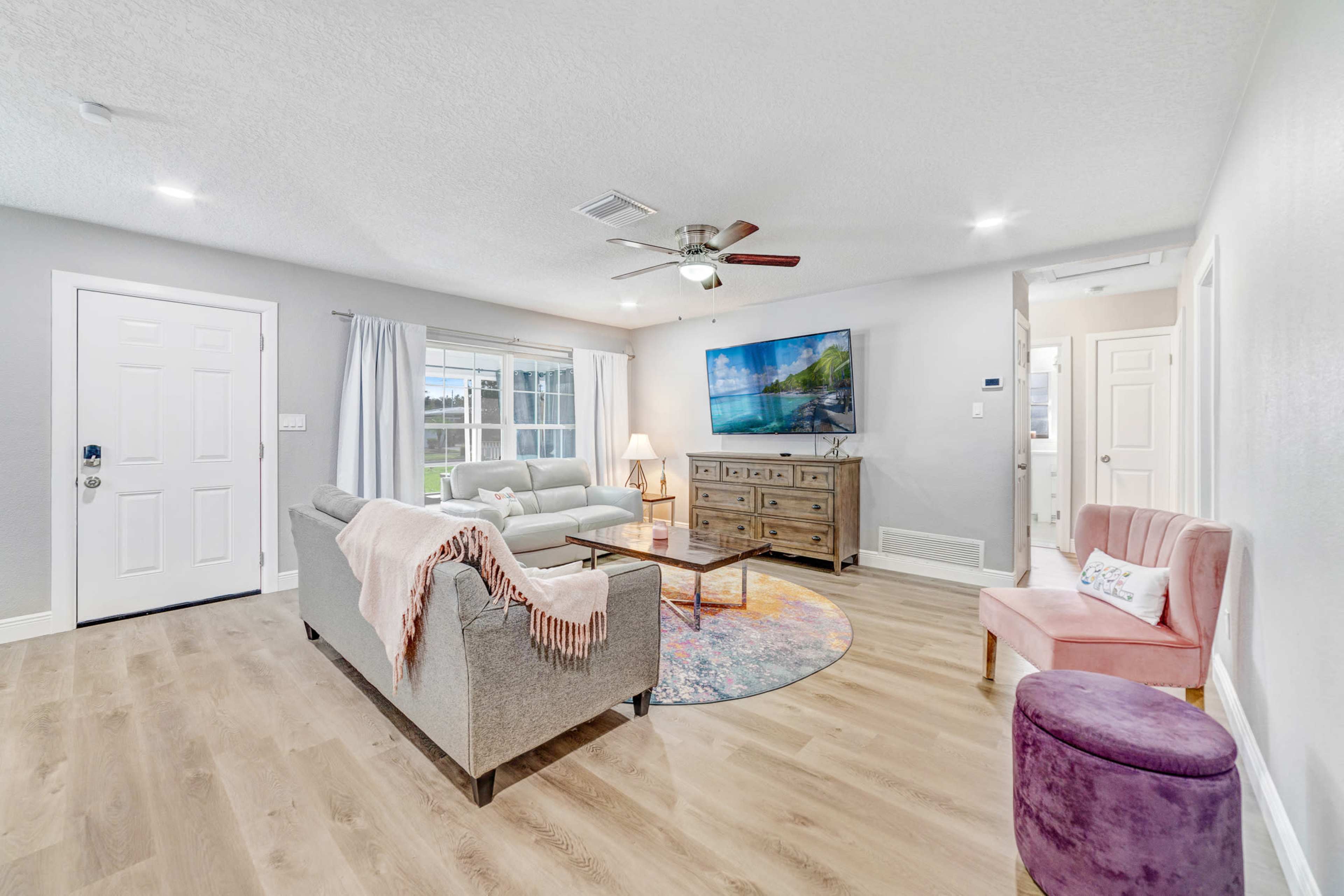 A modern living room features a light-colored sofa, a pink armchair, a wooden cabinet, and a large TV mounted on the wall, with a rug under a coffee table.