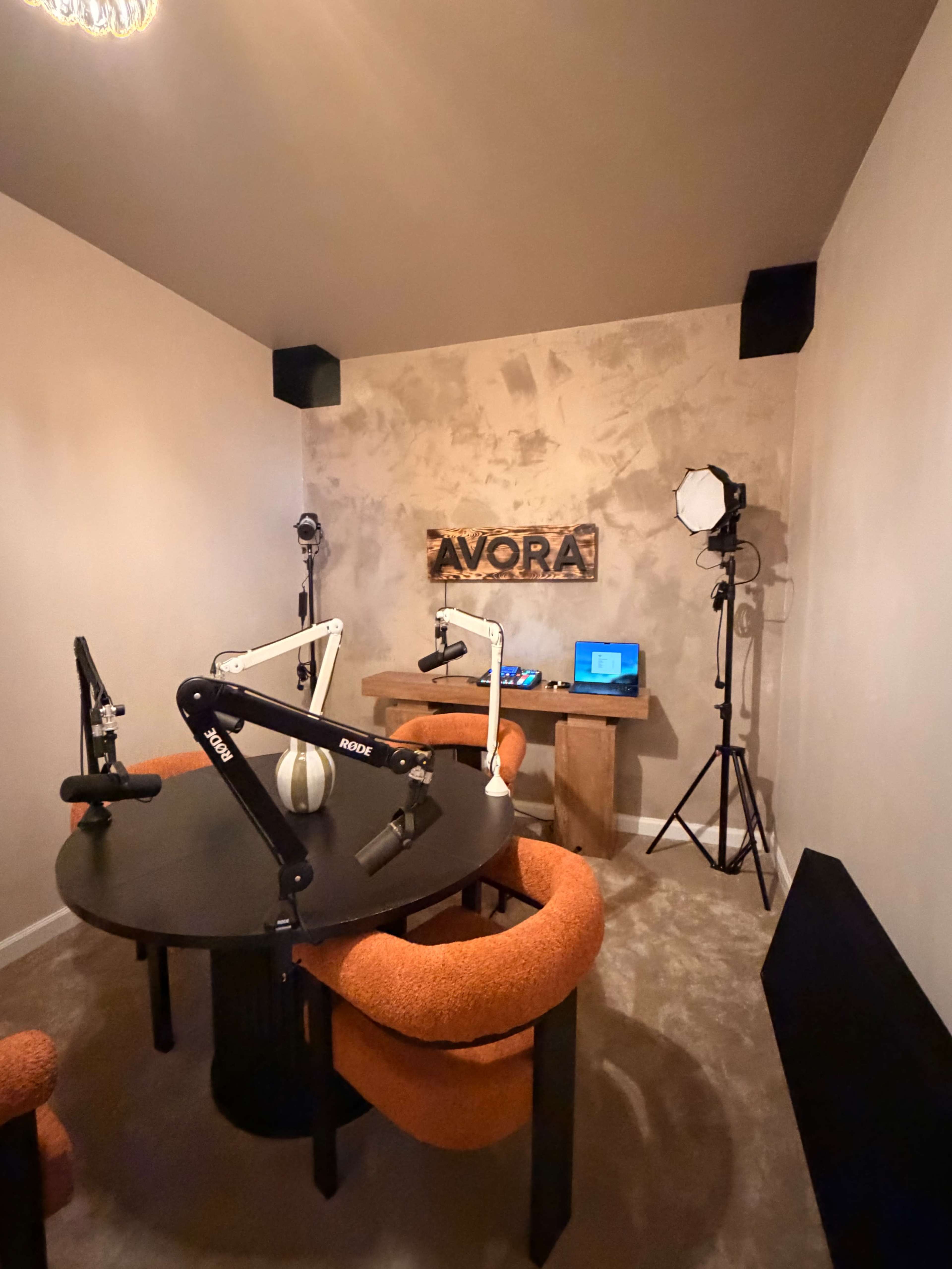 The image shows a small recording studio with a round black table, orange chairs, and equipment for podcasting or video recording.