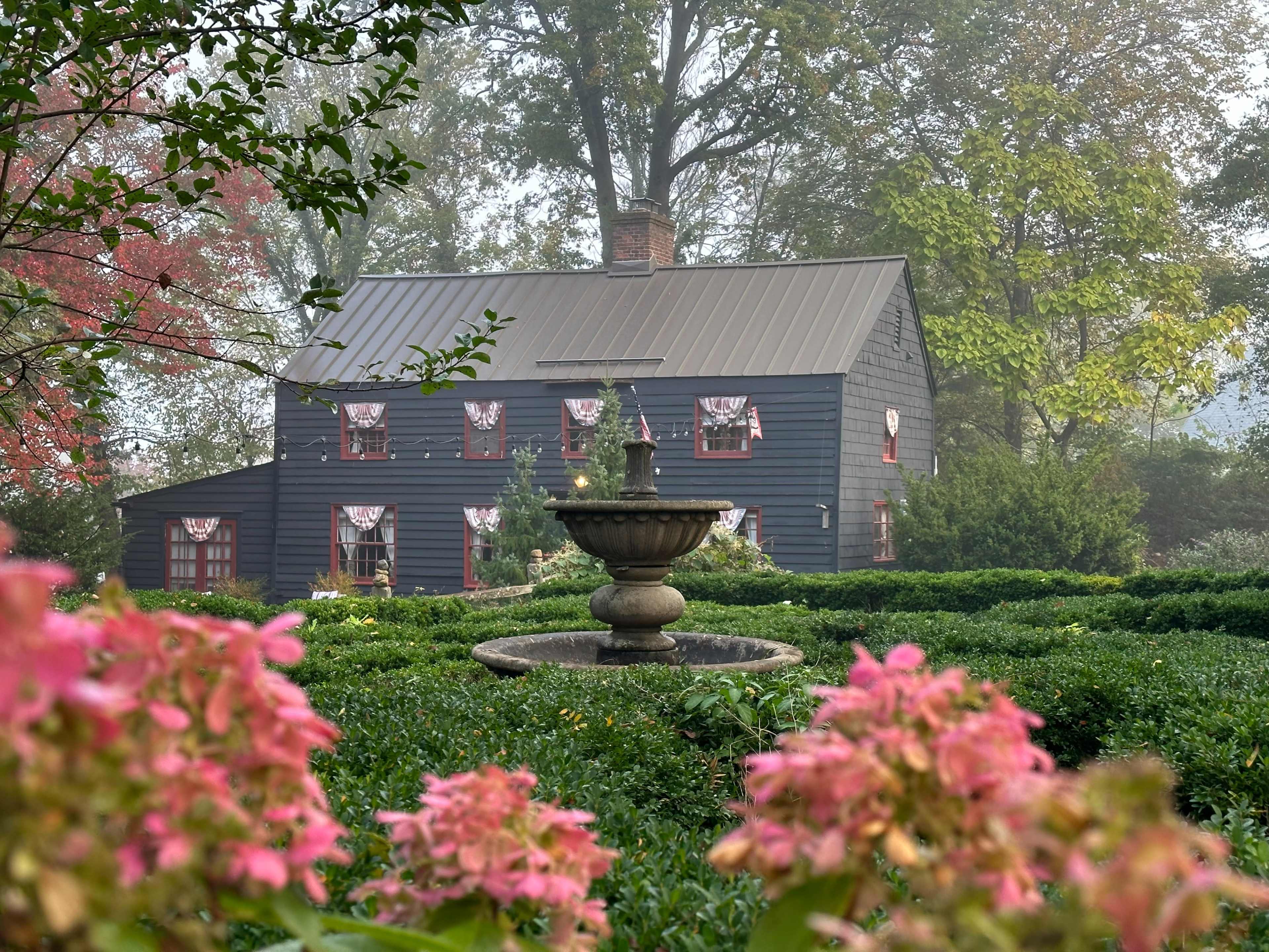 A dark blue house with a metal roof is surrounded by neatly trimmed hedges and a decorative fountain in a garden filled with pink flowers.