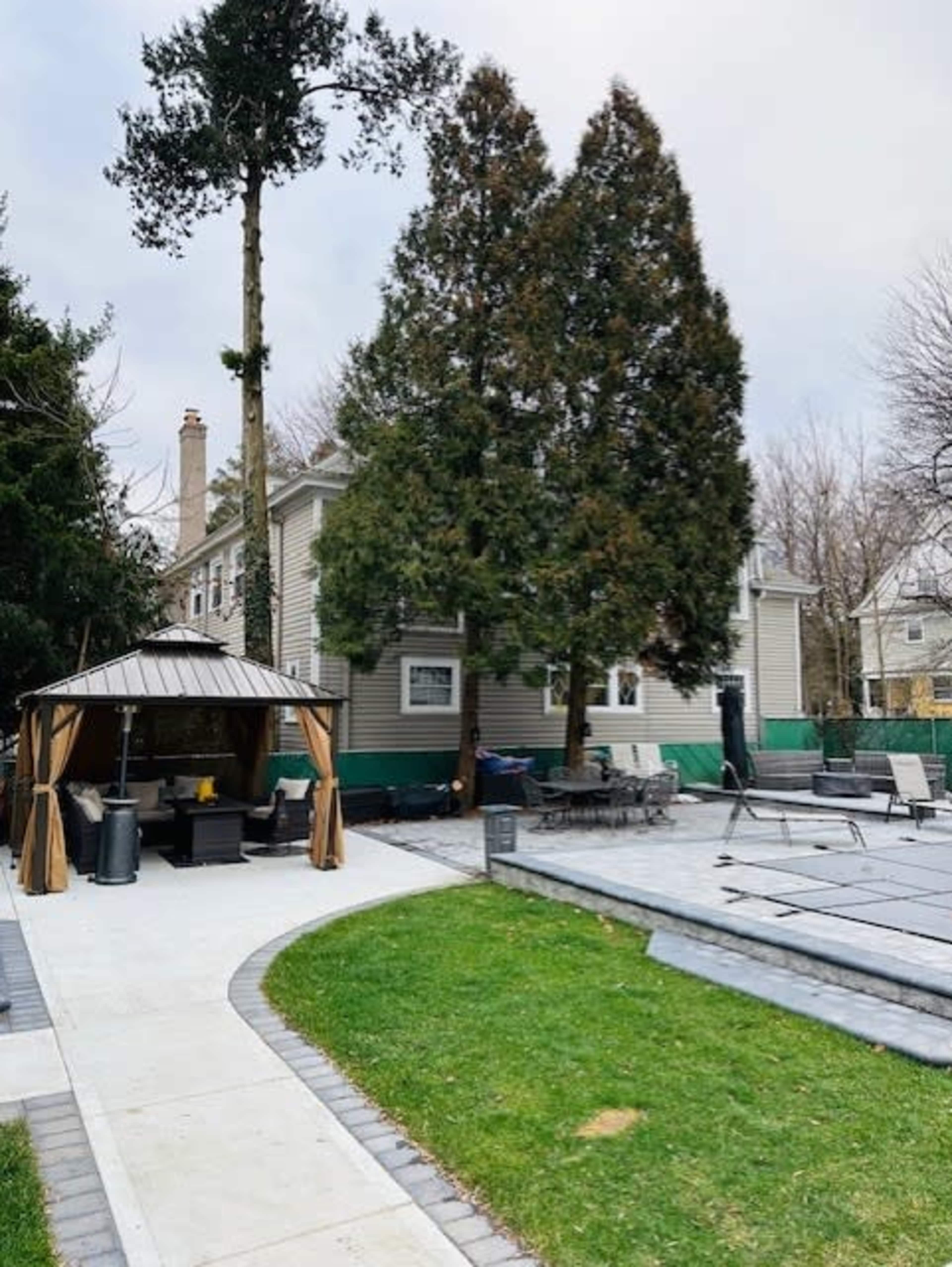A backyard with a gazebo, patio furniture, a paved walkway, and several large trees near a house.