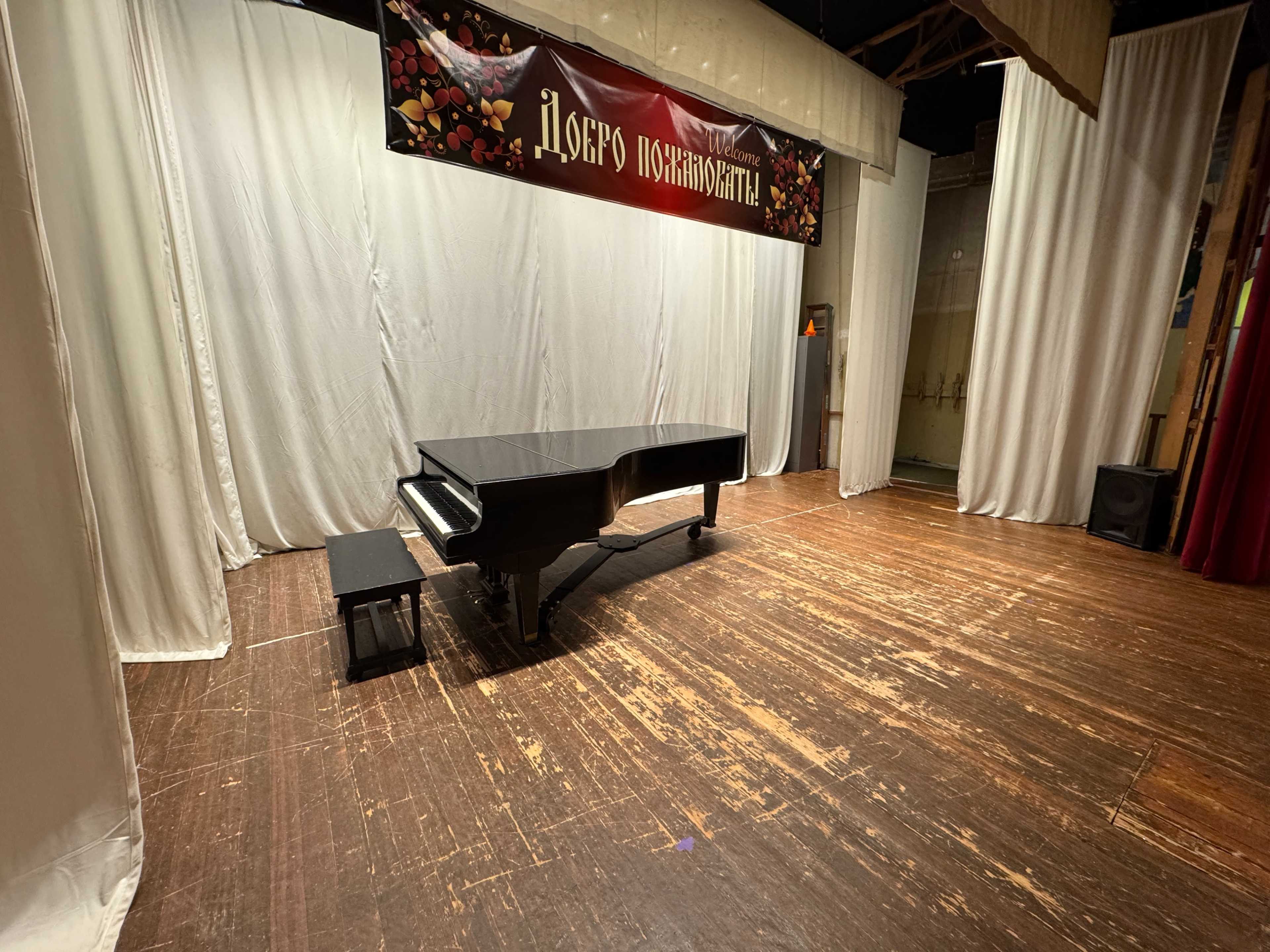 A black grand piano is placed on a wooden stage covered with a light-colored curtain backdrop and a decorative banner.