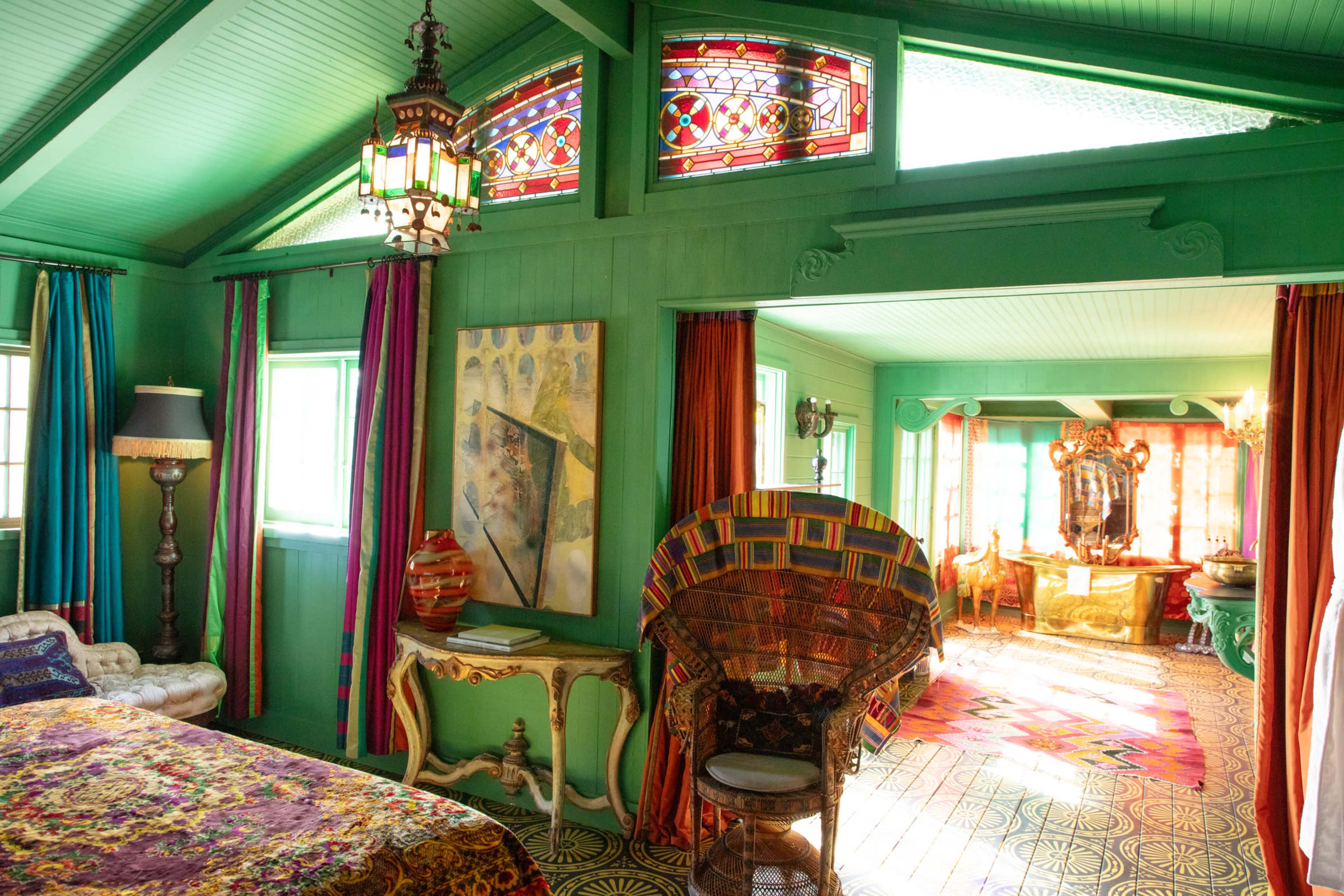The image shows a vibrantly decorated room with green walls, colorful curtains, and stained glass windows, featuring a bed, artwork, and unique furniture.