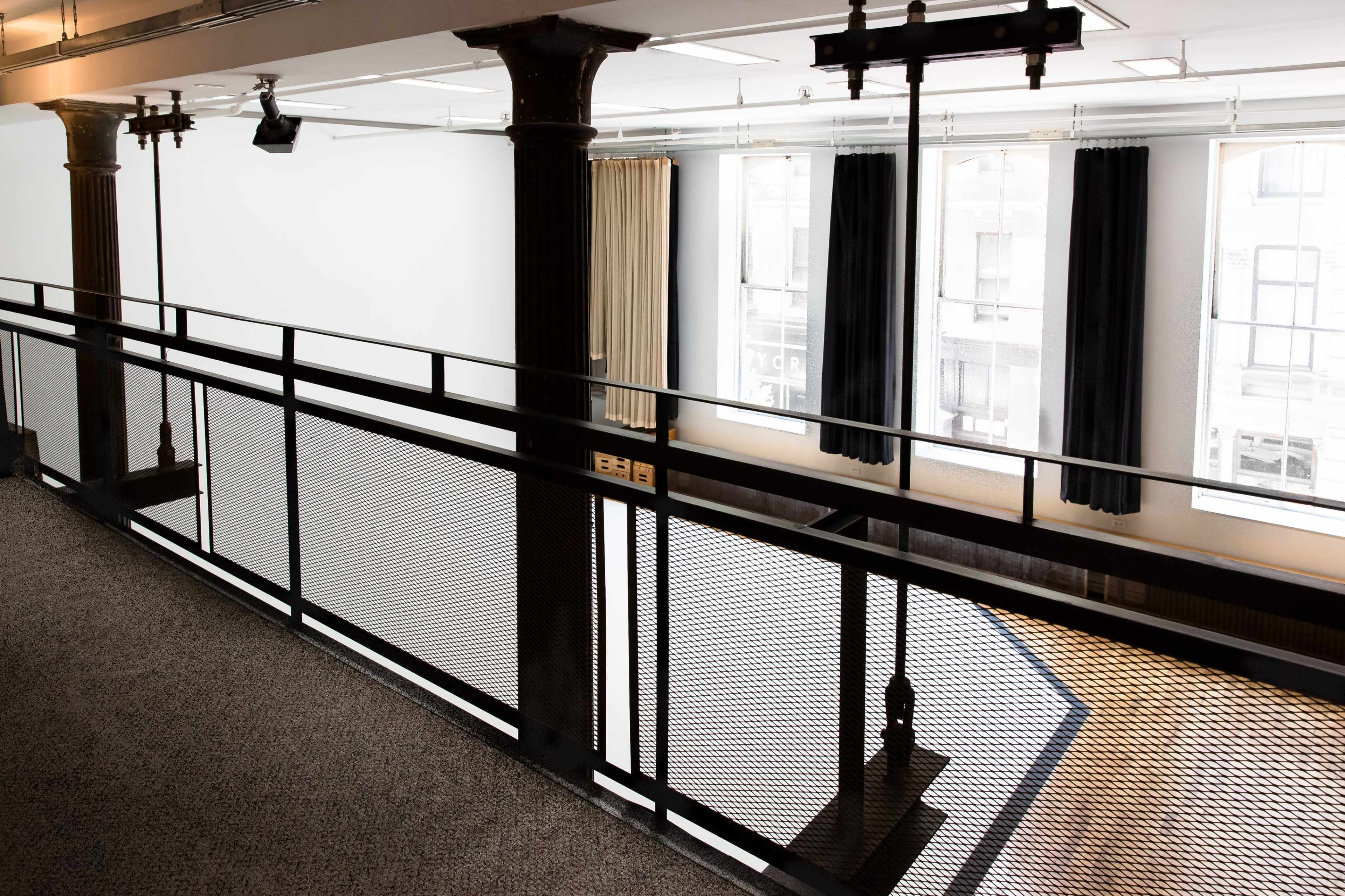 A balcony with a metal railing overlooks a bright, empty room with large windows and dark drapes.