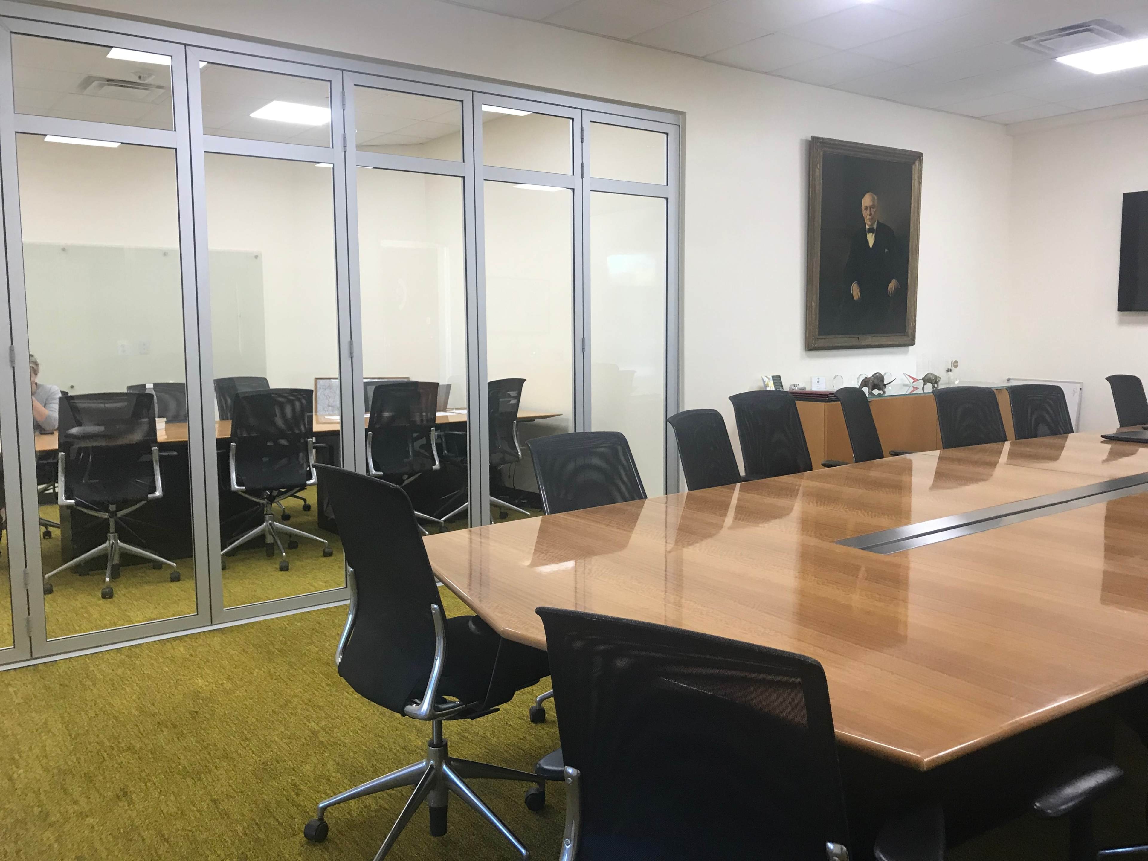 A large conference room features a wooden table surrounded by black chairs, with glass walls enclosing an adjacent meeting space and a portrait hanging on the wall.