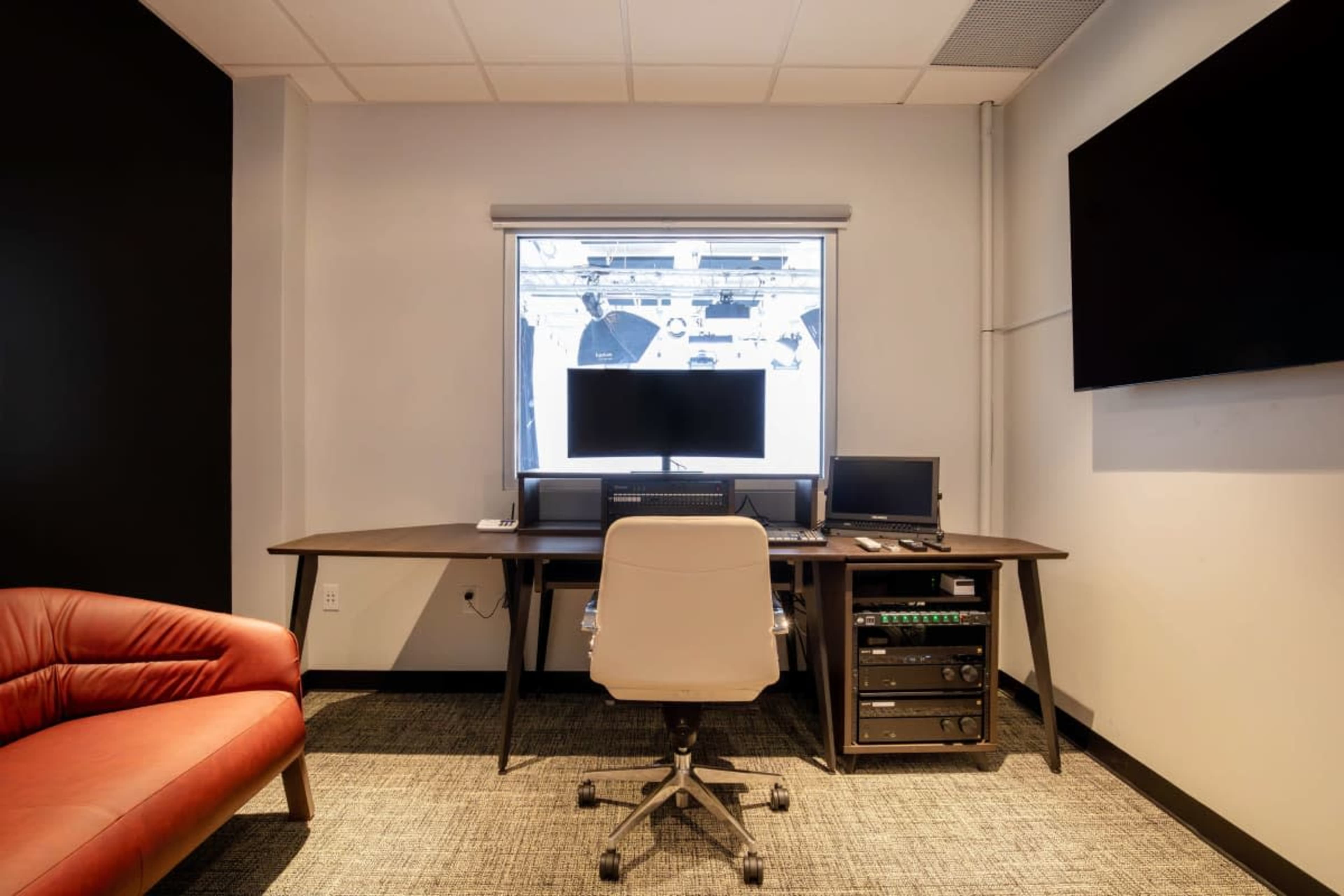 The image shows a modern office space with a dark wall, a window overlooking a studio setup, a desk with two computer monitors, and a red leather chair.