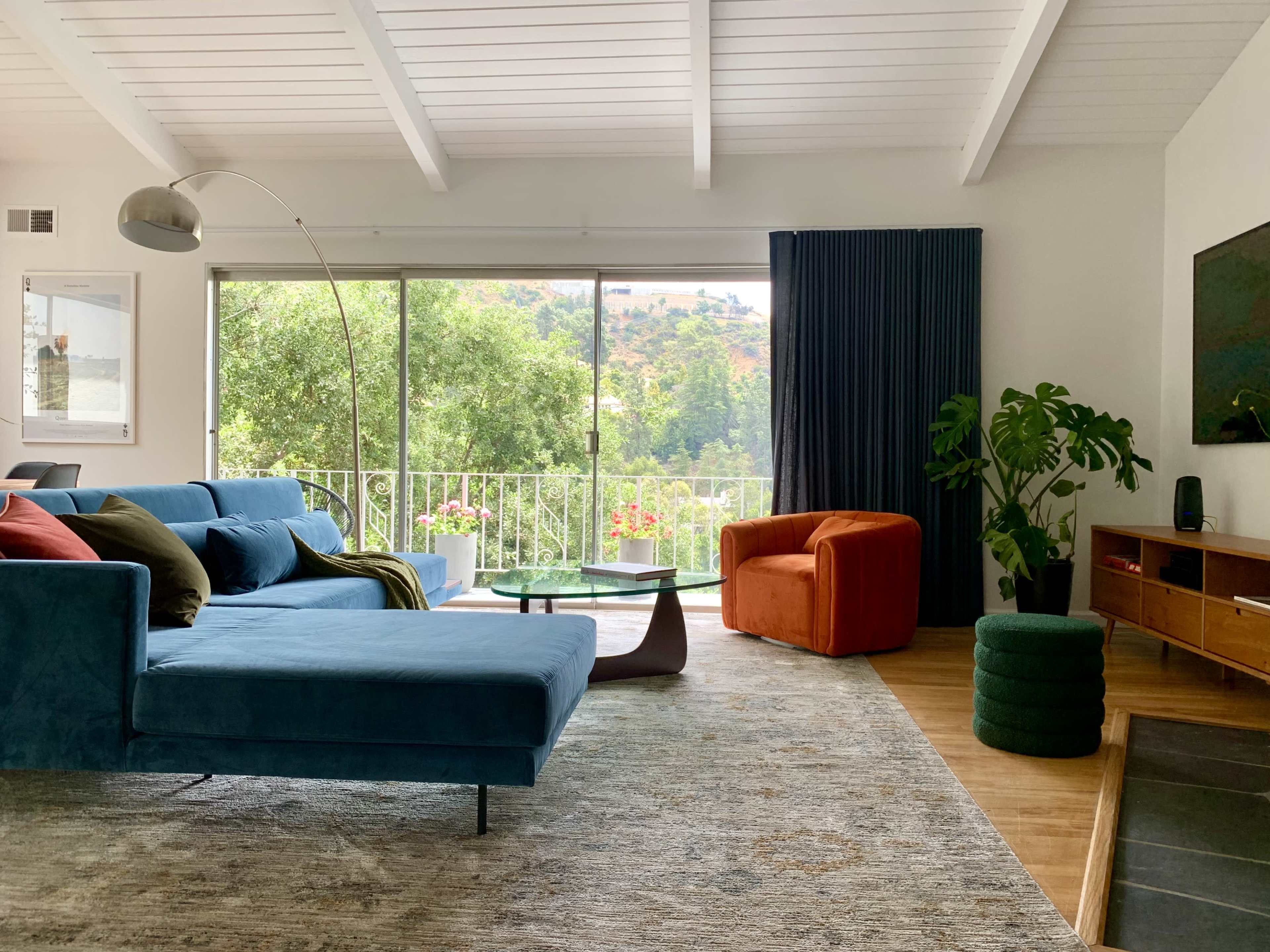 The interior scene features a modern living room with a blue sectional sofa, an orange chair, large windows showcasing greenery, and a mix of decorative plants and furniture.