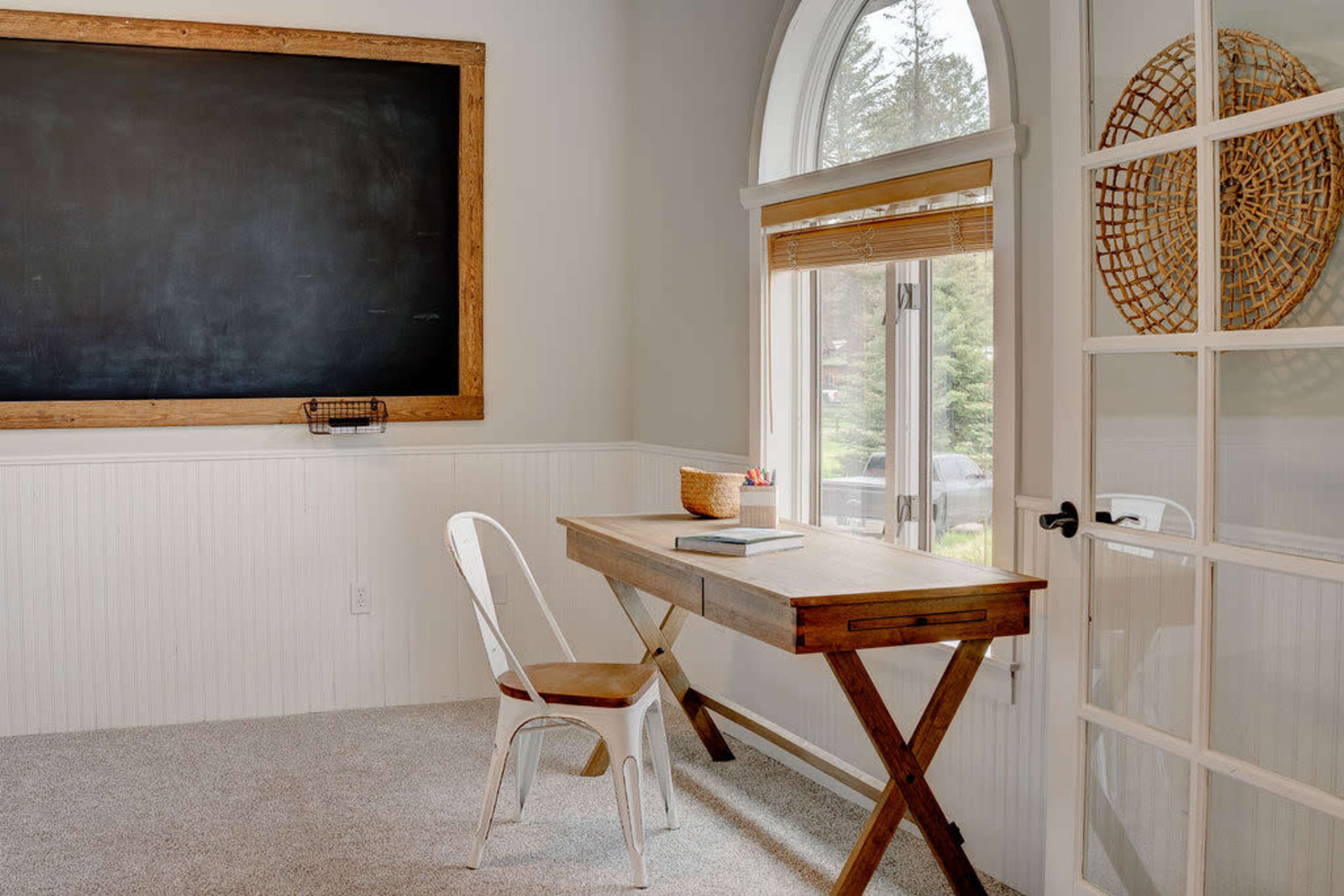 A minimalist home office with a wooden desk, a white chair, a chalkboard on the wall, and a large window with natural light.