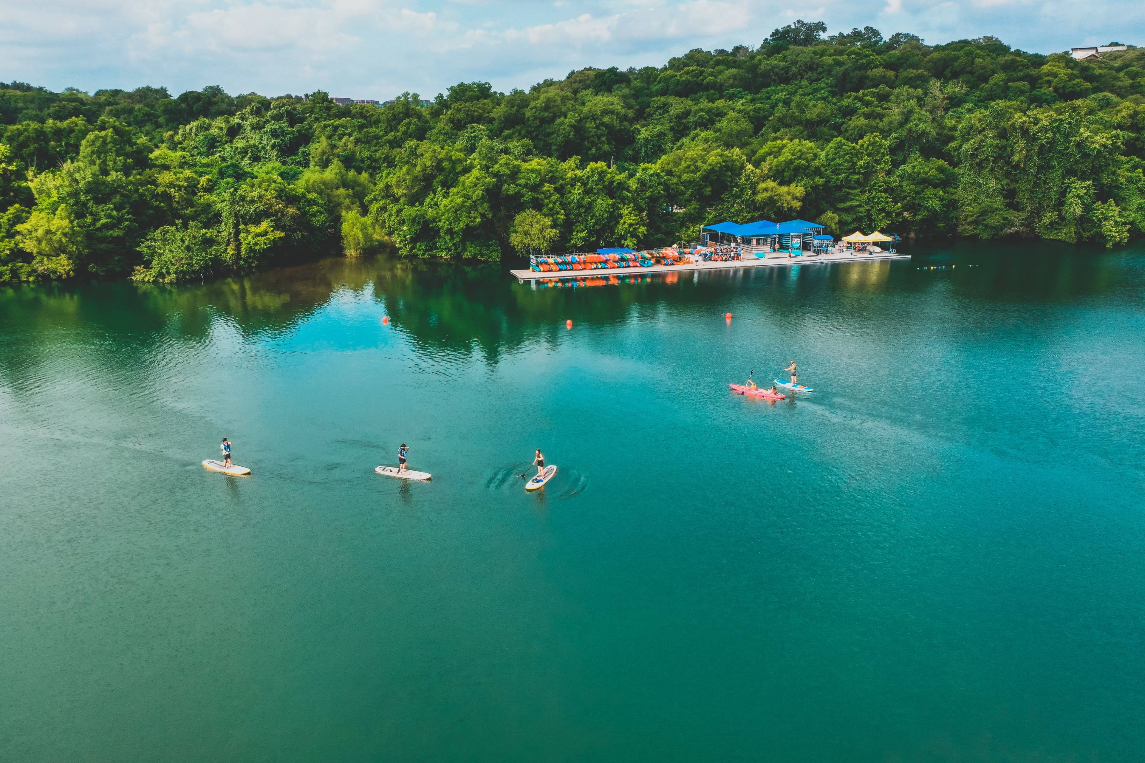 The image shows several people paddleboarding on a calm blue lake, with a backdrop of lush green trees and a small resort area featuring colorful umbrellas and boats.