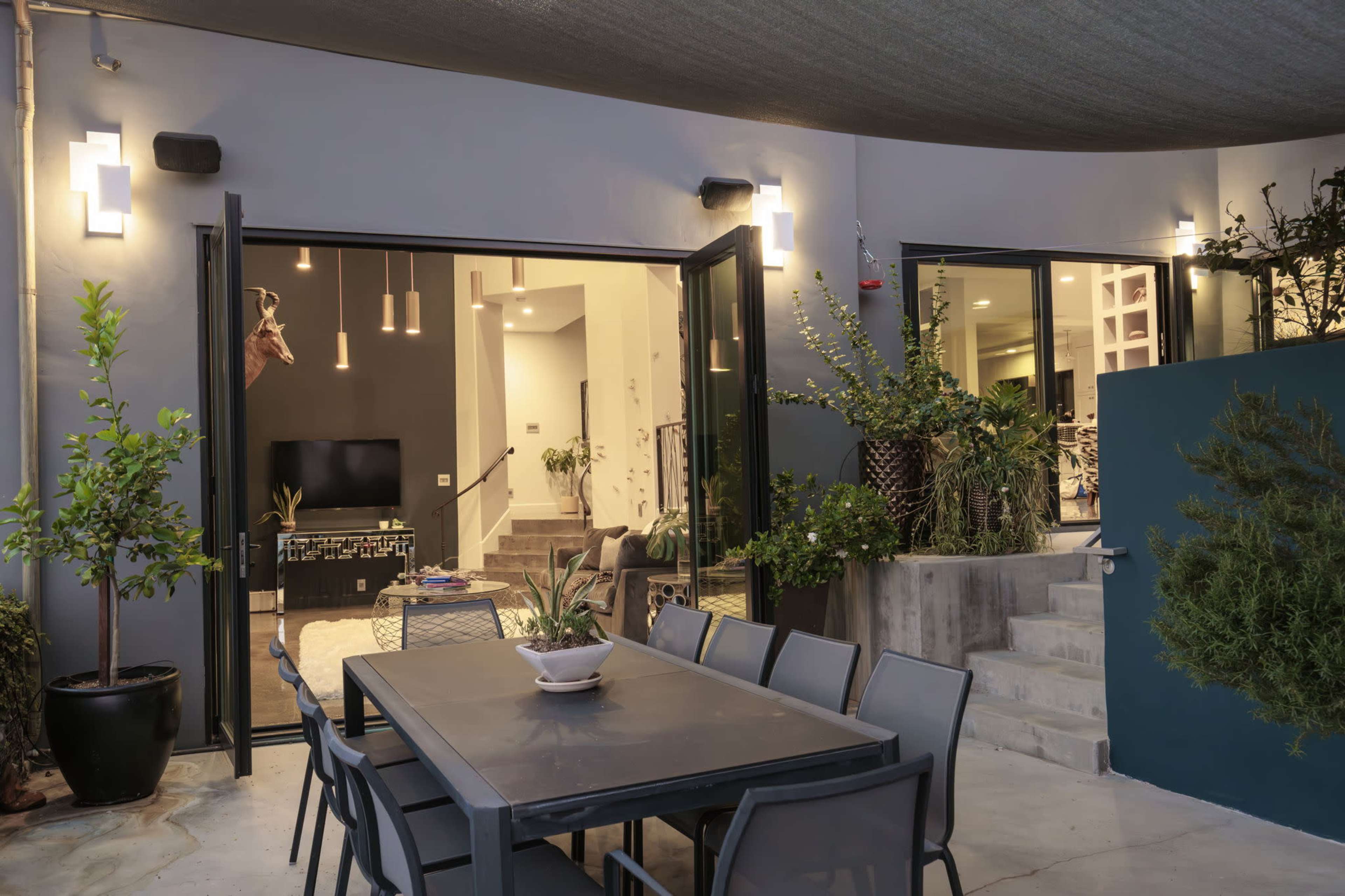 A patio area features a dining table surrounded by chairs, with open doors leading into an interior space decorated with plants and modern furnishings.