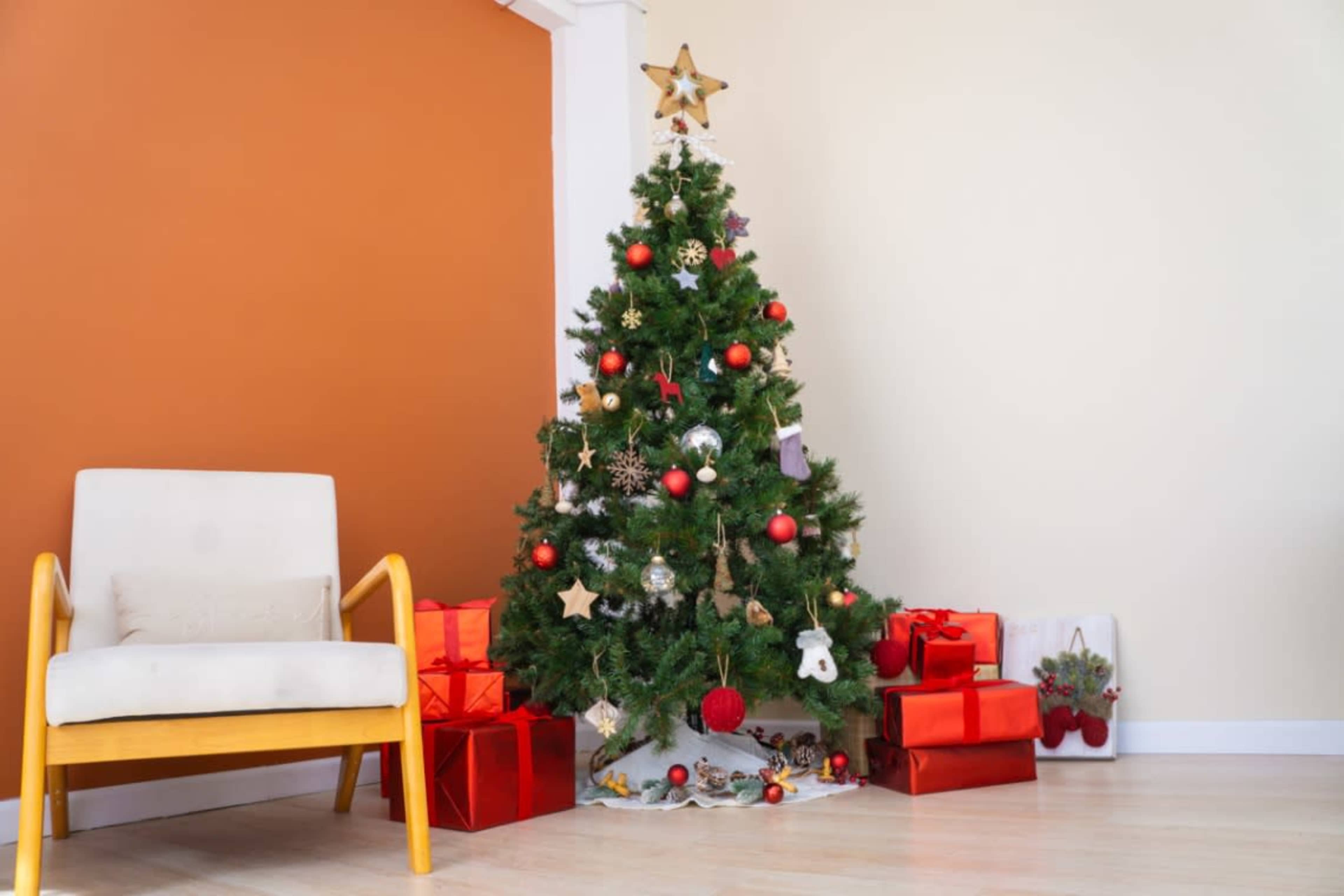 A decorated Christmas tree stands in the corner of a room beside a light-colored chair and several wrapped gifts.