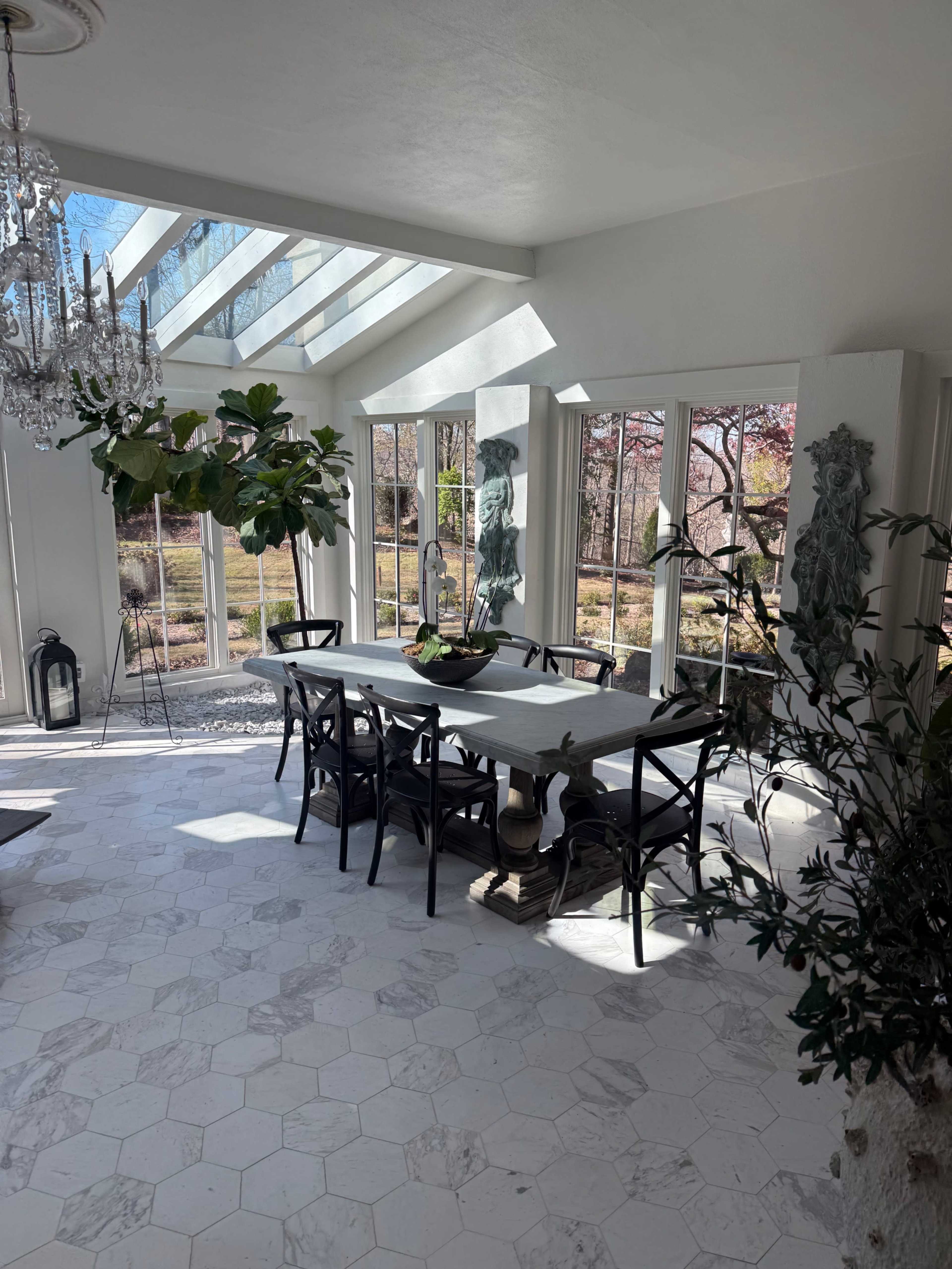 A sunlit dining room features a large table surrounded by chairs, with large windows and decorative plants framing the space.