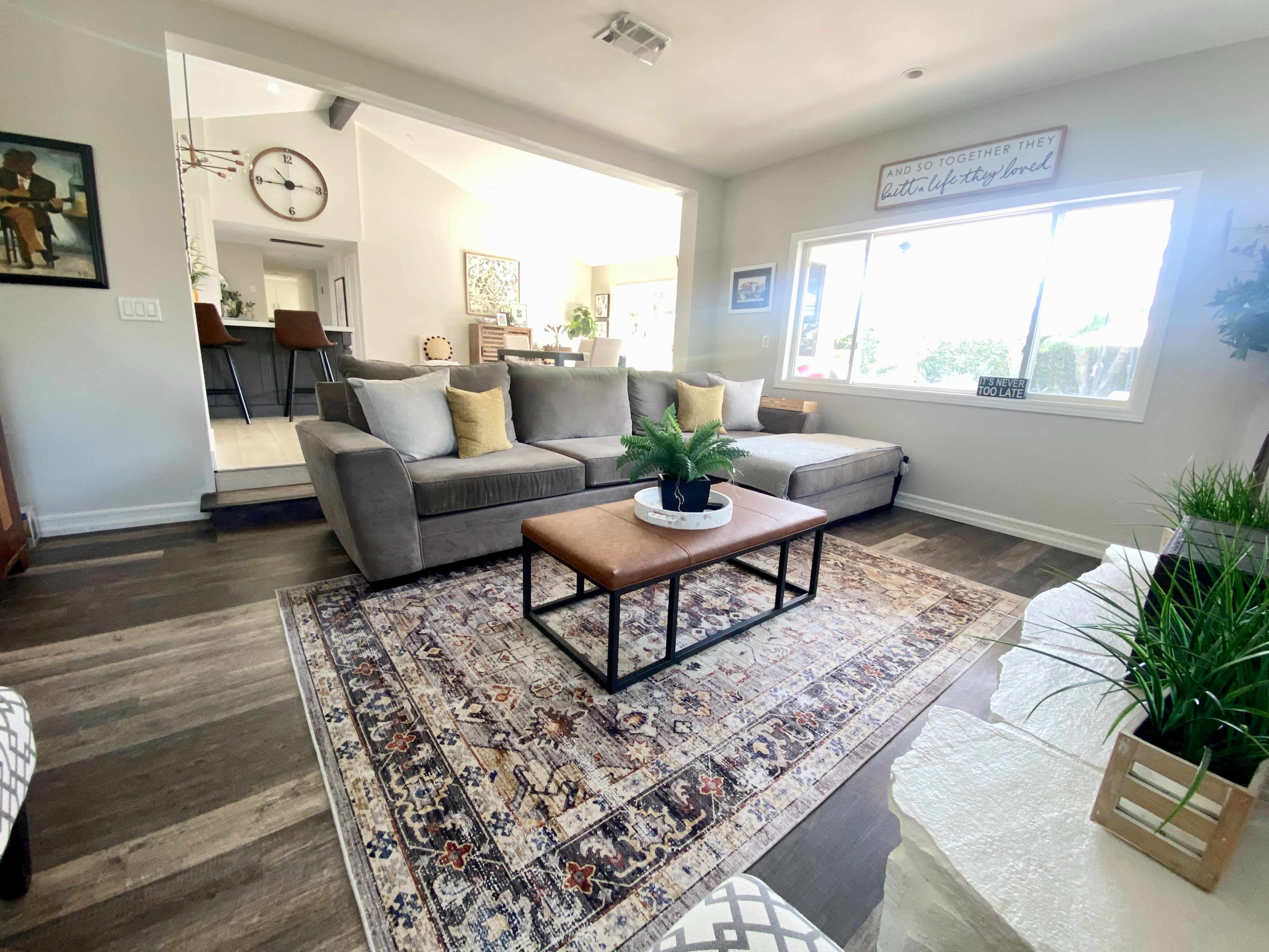 The image shows a living room with a gray sofa, an orange coffee table on a patterned rug, and windows that allow natural light to enter the space.