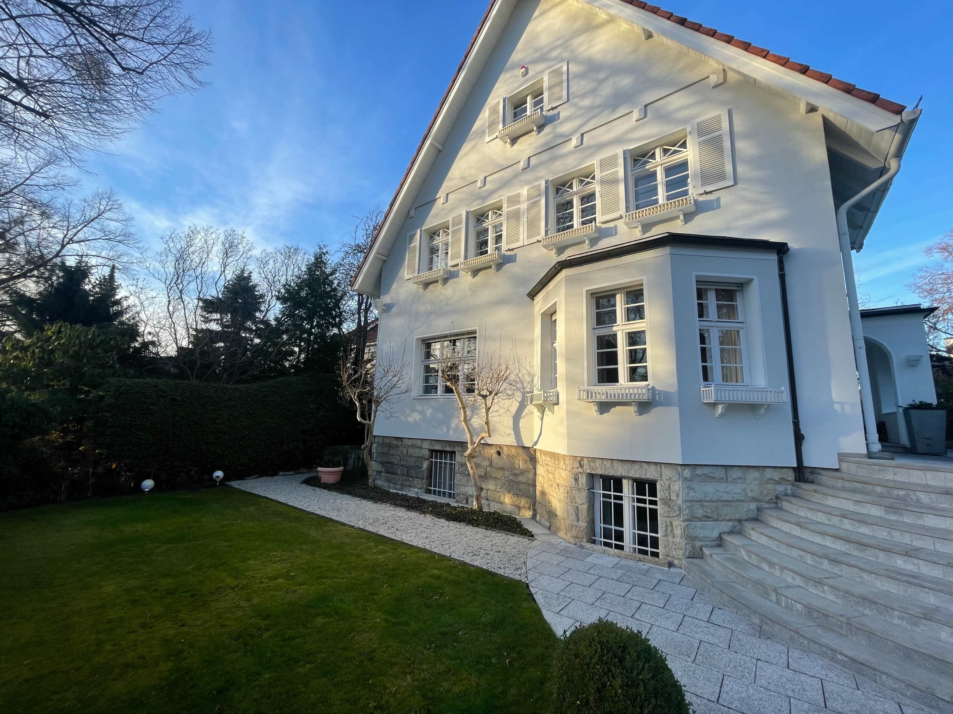 A large, white, three-story house with multiple windows, surrounded by a well-maintained garden and stone steps leading to the entrance.
