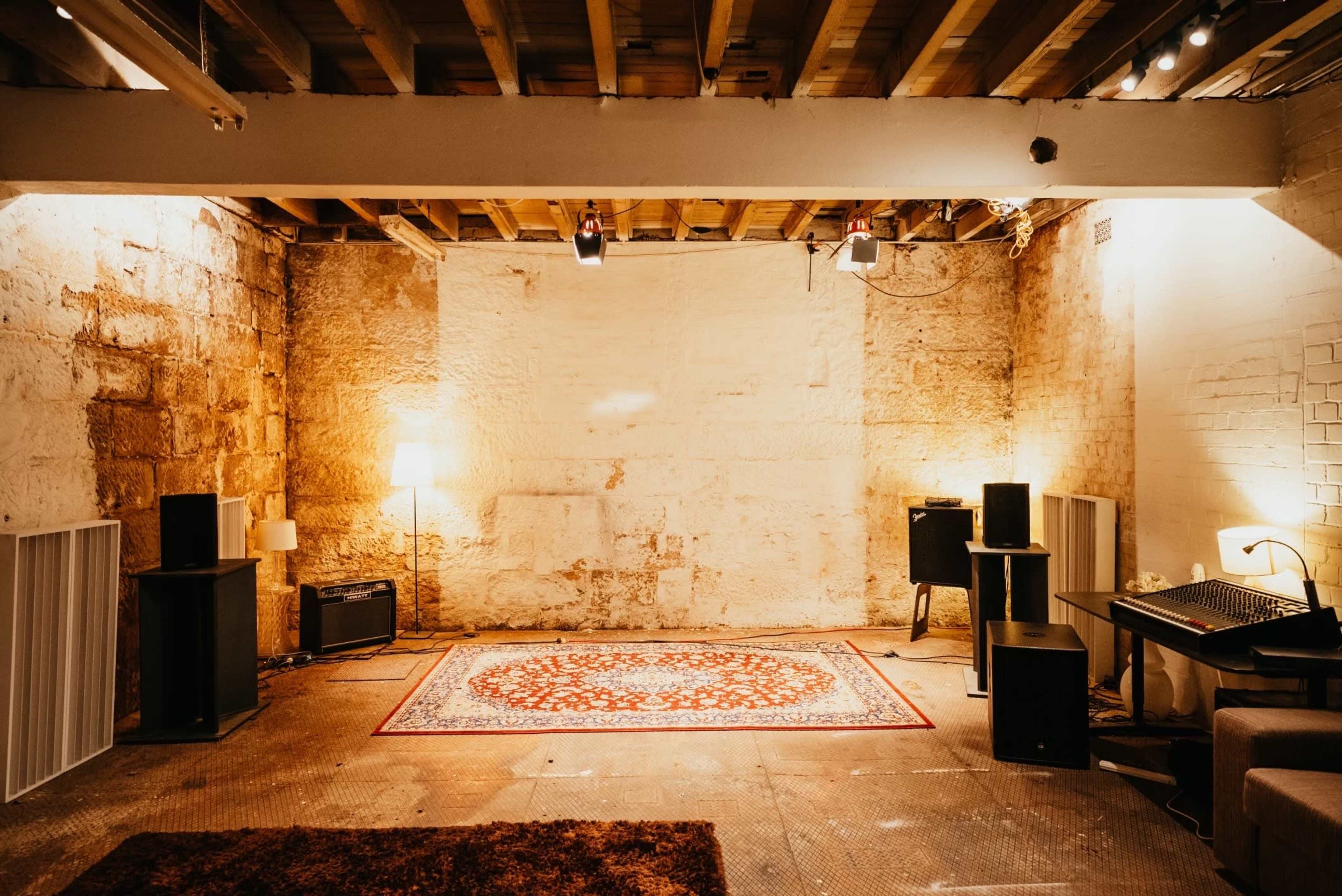 A spacious room with exposed stone walls features a central rug, speakers, and a desk with audio equipment, illuminated by soft lighting.