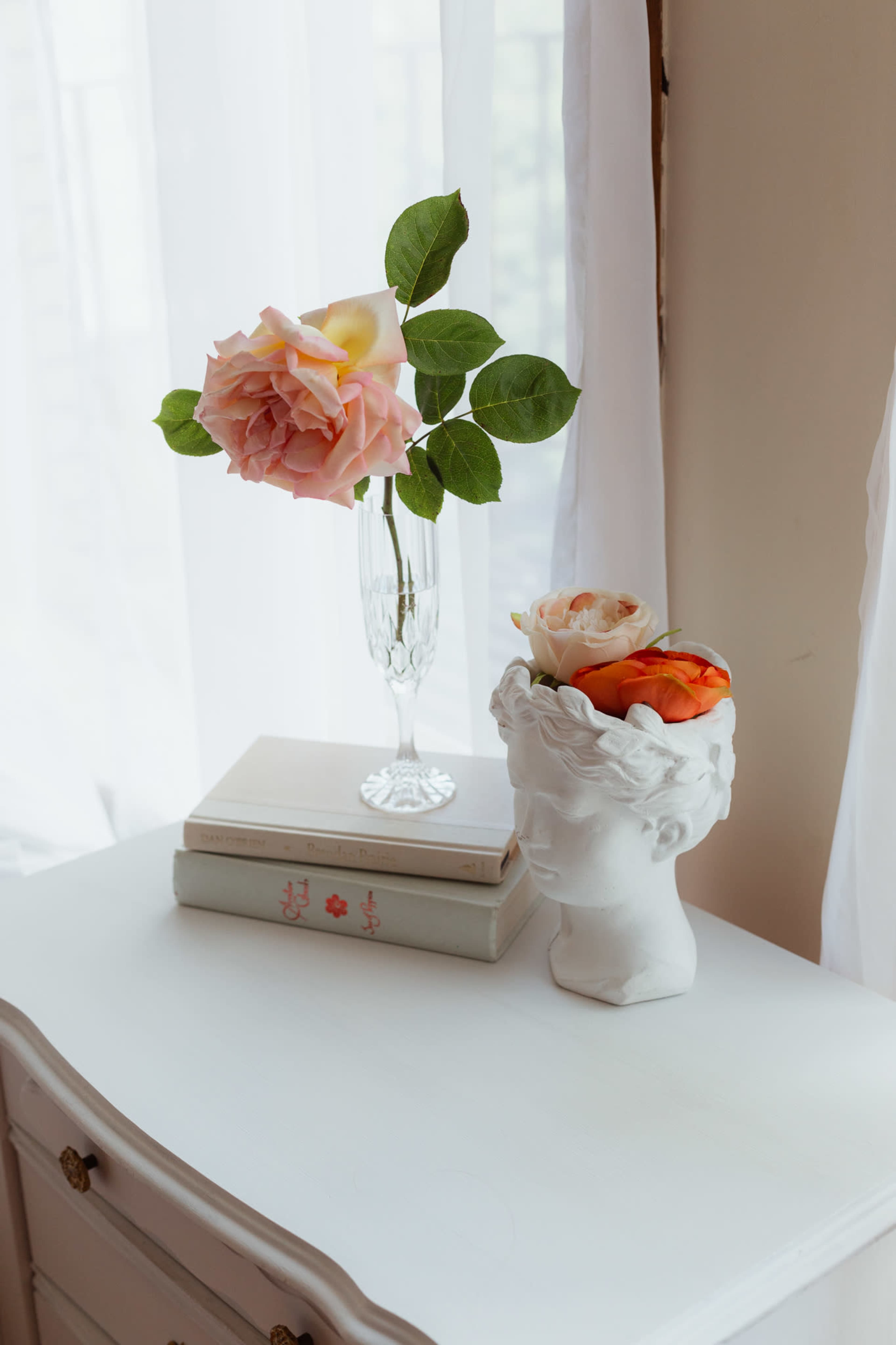 A clear vase with pink roses and green leaves sits on top of stacked books next to a decorative head-shaped vase containing orange flowers on a white dresser.