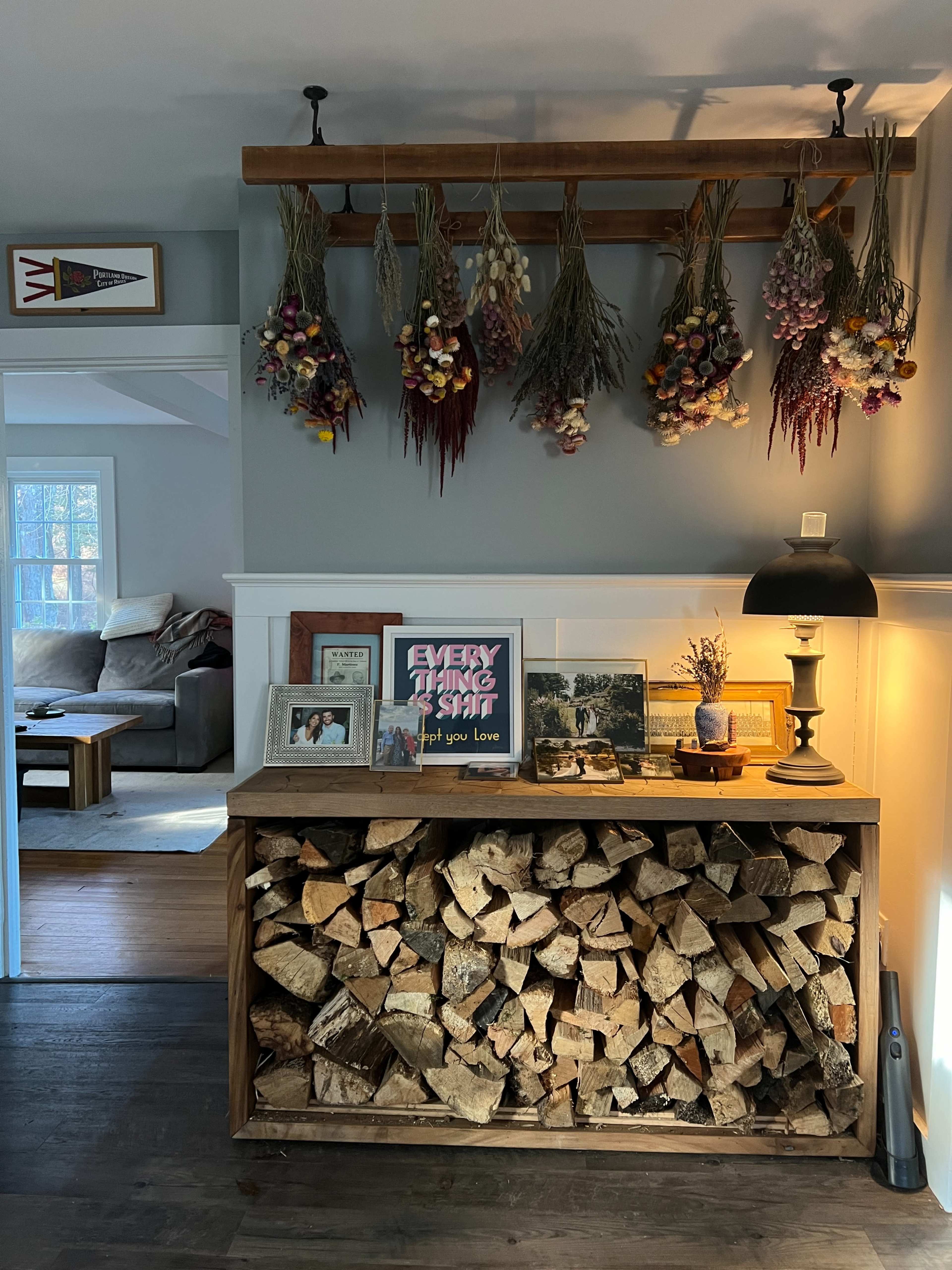 The image shows a cozy interior featuring a wooden storage unit stacked with logs, a decorative lamp, and dried flowers hanging above it, with a living area visible in the background.
