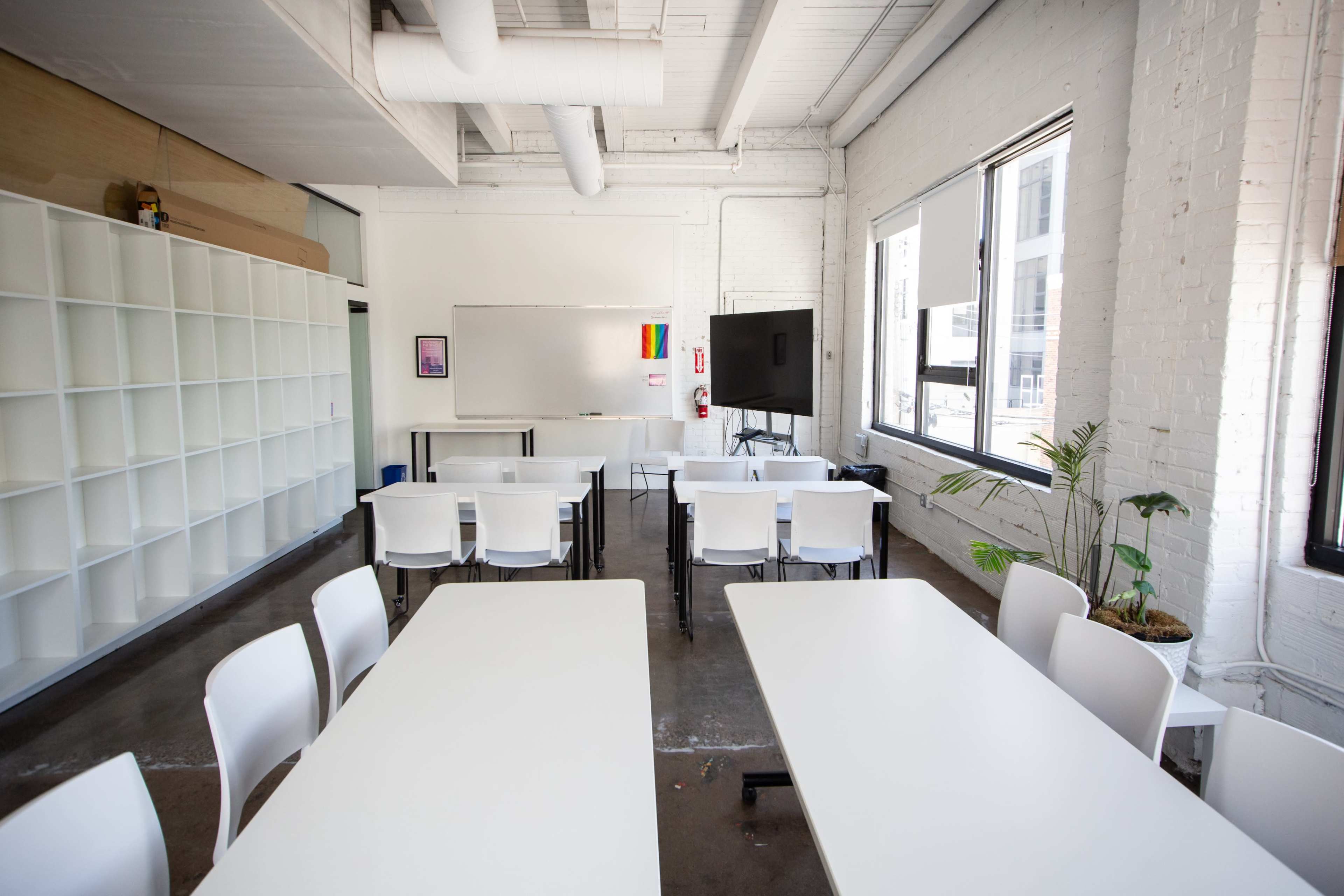 A brightly lit classroom features white tables and chairs arranged in rows, with a whiteboard and a large screen at the front, and shelving on one side.