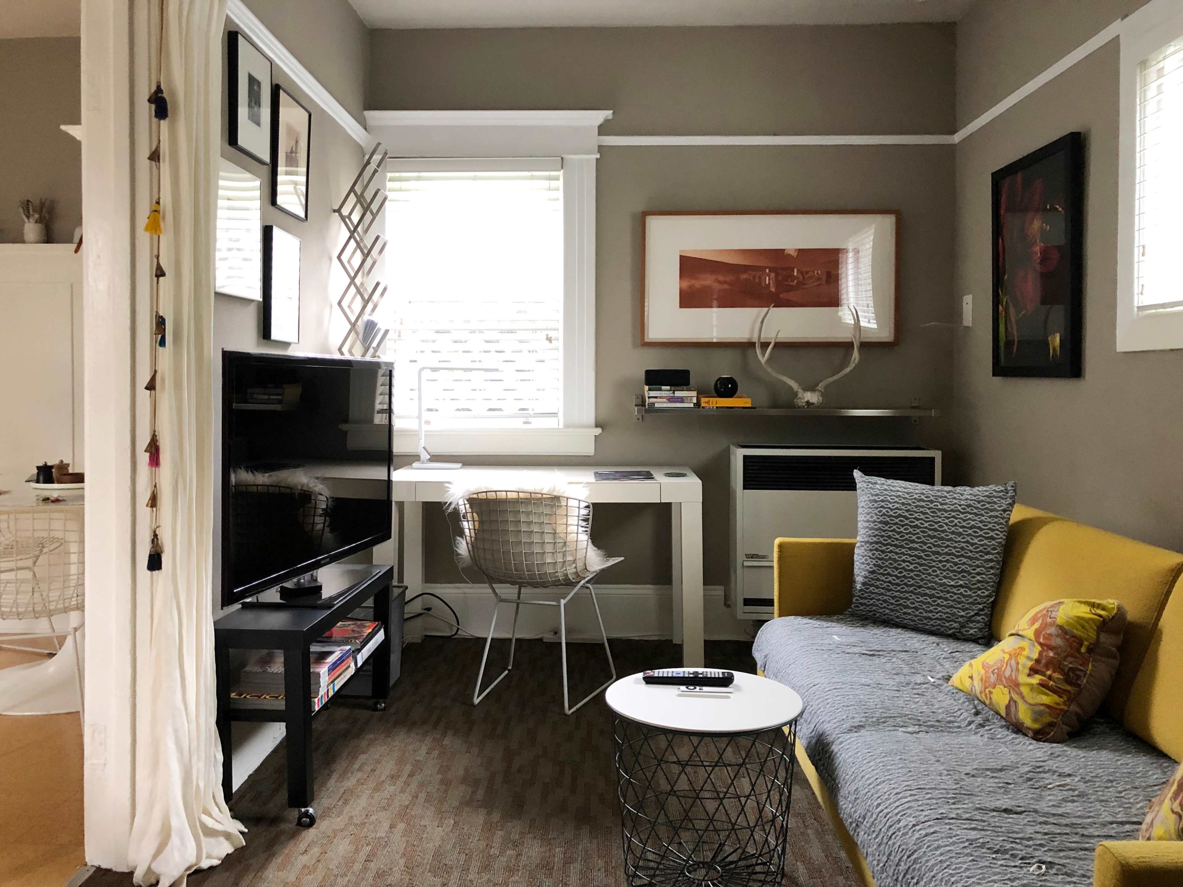 A small living area featuring a yellow sofa, a black TV stand, a white desk with a chair, and various framed artworks on the walls.