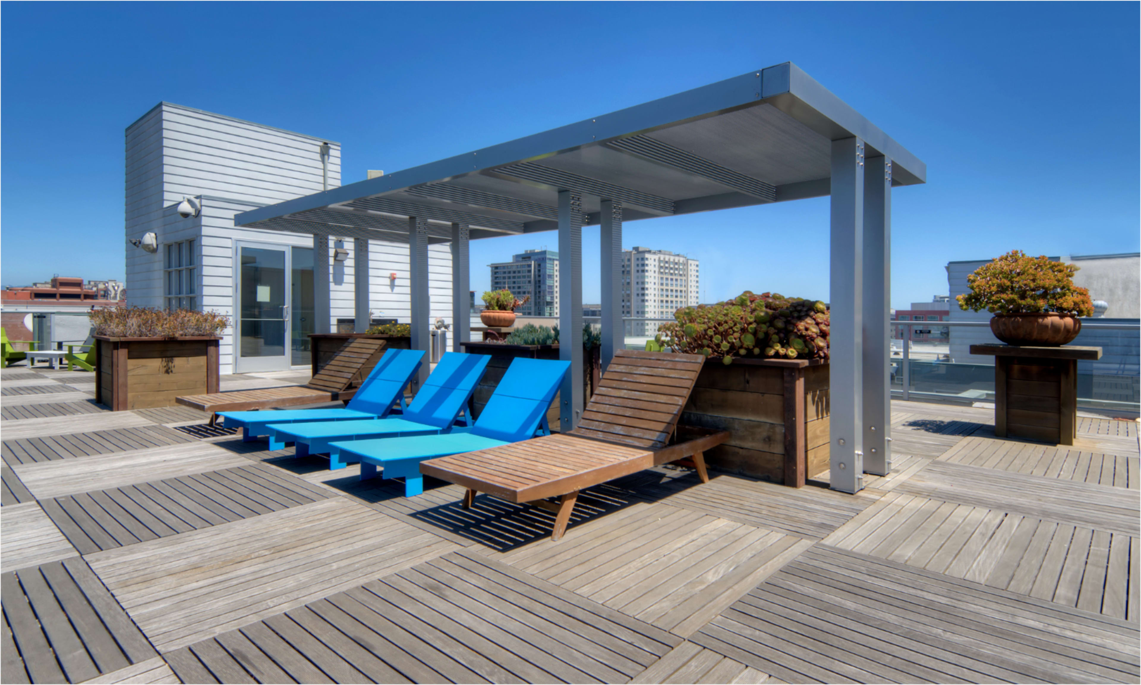 The image shows a rooftop deck featuring blue loungers, wooden seating, and planters under a metal canopy with a clear blue sky above.