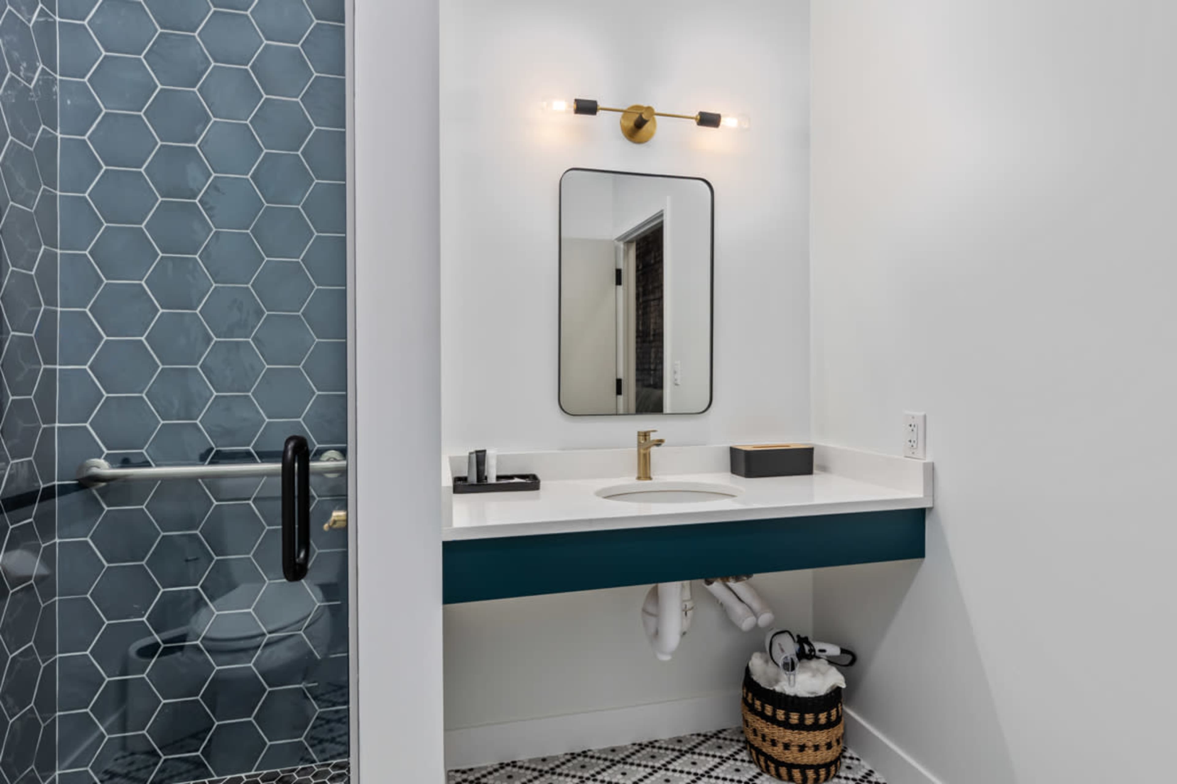 A modern bathroom featuring a hexagonal tiled shower, a wall-mounted mirror, a floating sink with a gold faucet, and a patterned floor.