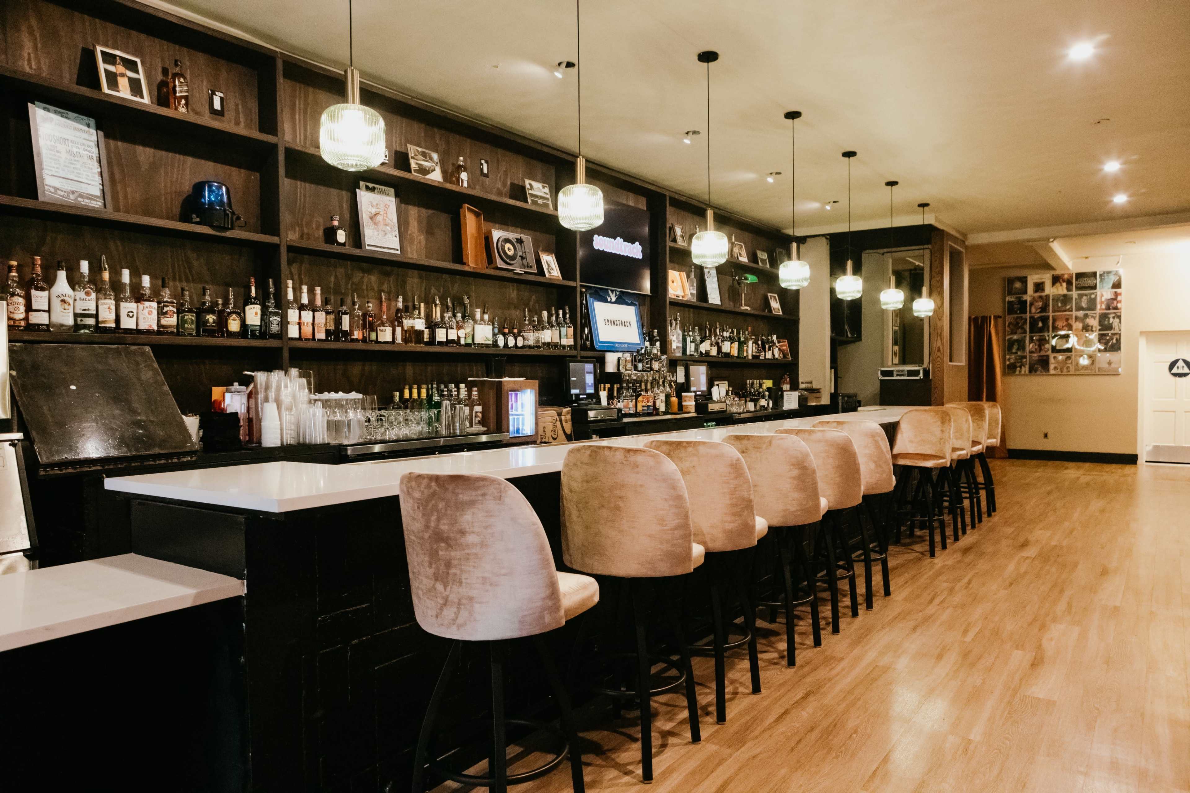 The image shows a modern bar with a long counter and high-backed chairs, decorated with shelves of various bottles and illuminated by hanging lights.