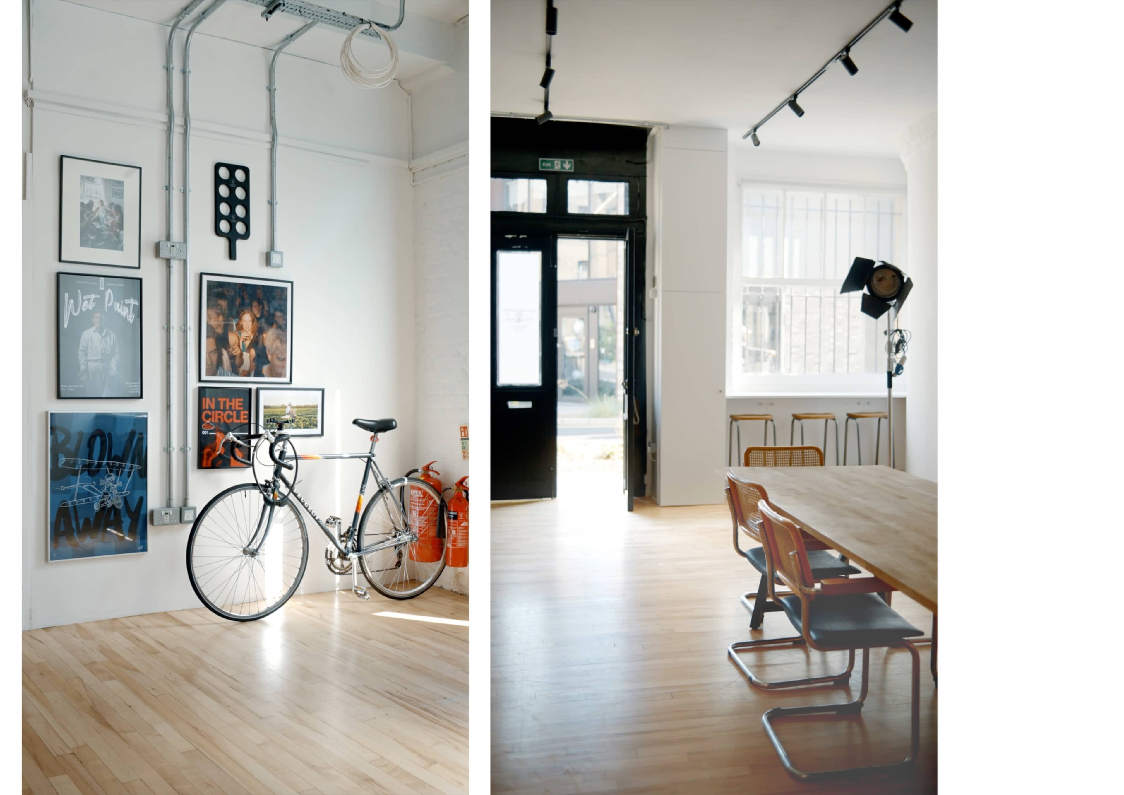 A modern interior space featuring a bicycle against a wall with framed art on one side, and a dining area with a large wooden table and metal chairs on the other.