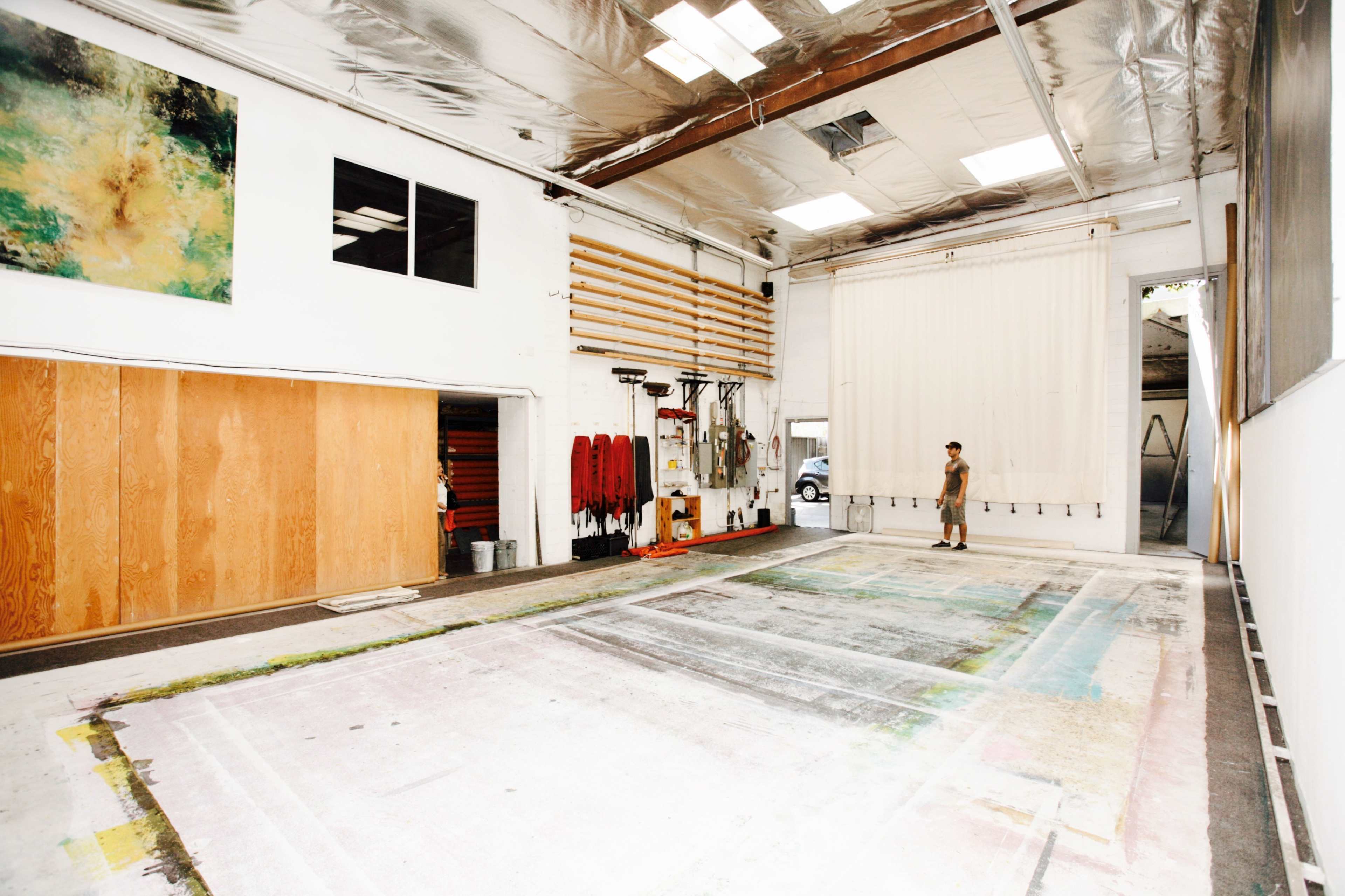 A person stands in an expansive, well-lit studio space with a large, covered floor and several art supplies and tools stored along the walls.