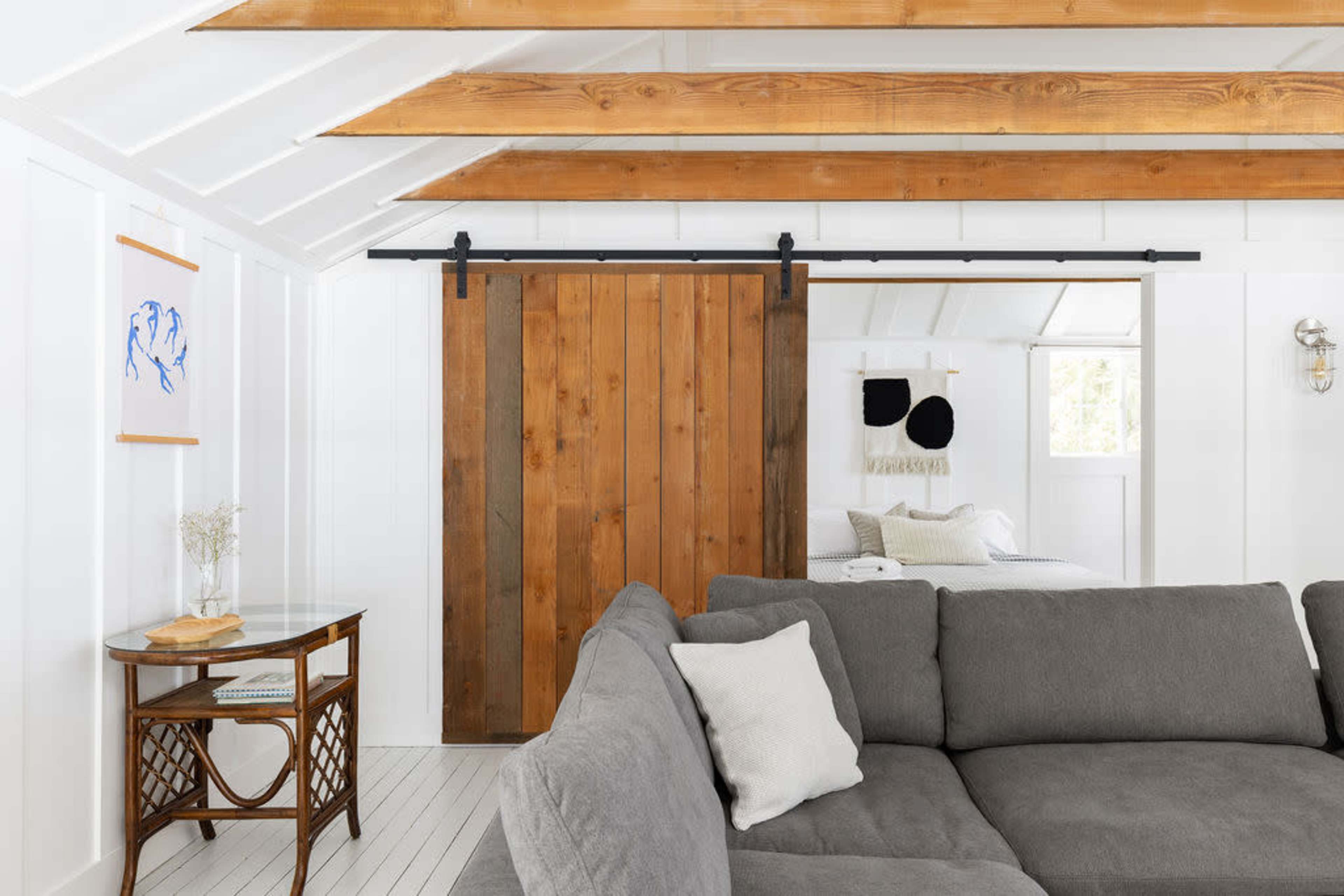 The image shows a cozy interior with a gray sectional sofa facing a wooden sliding barn door, which leads to a bright space with a bed and decorative wall art.