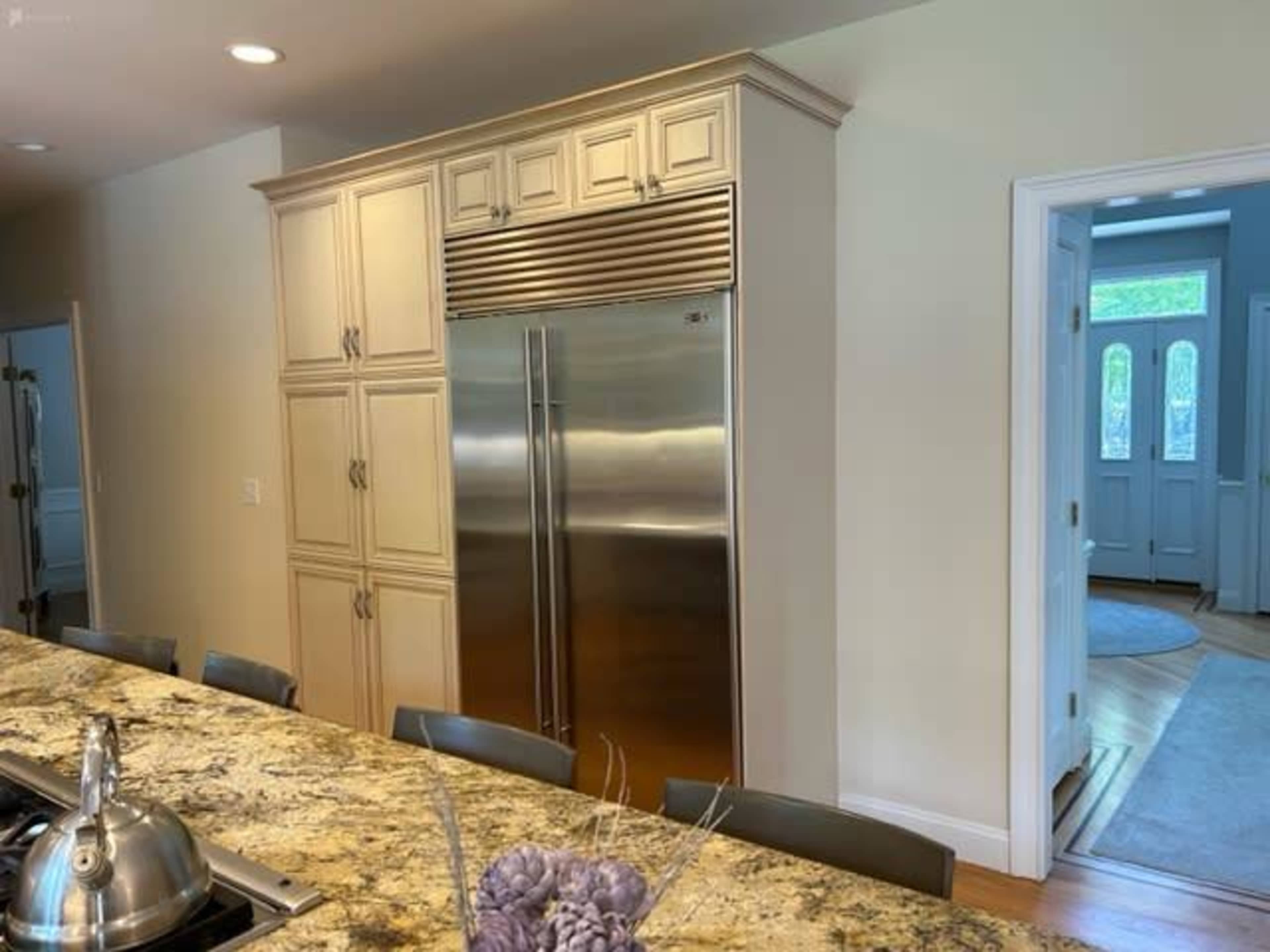 The image shows a modern kitchen with a stainless steel refrigerator integrated into light-colored cabinetry beside a marble countertop and a doorway leading to another room.