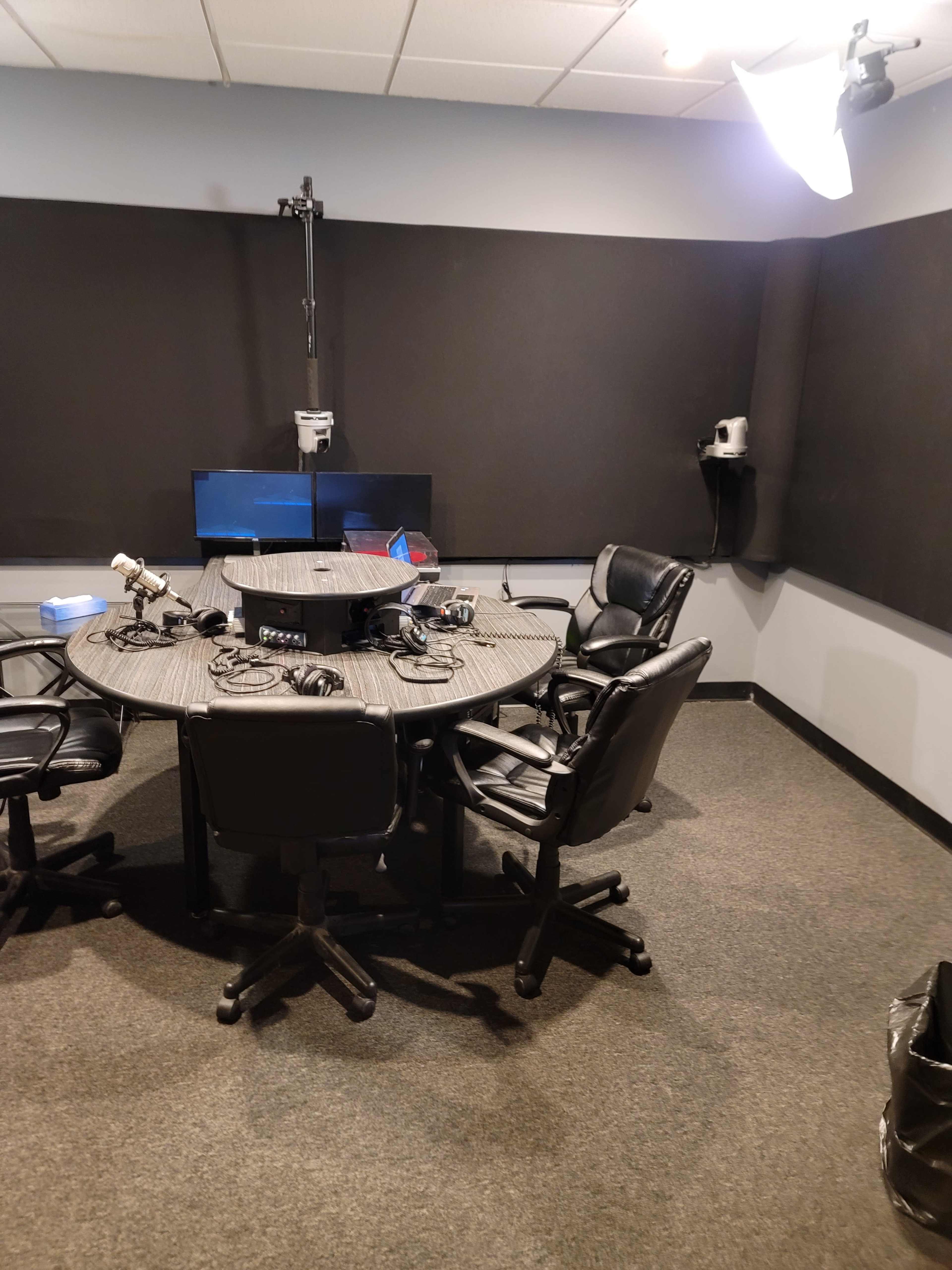 A soundproof recording studio with a round table, five black chairs, and audio equipment set up for recording.