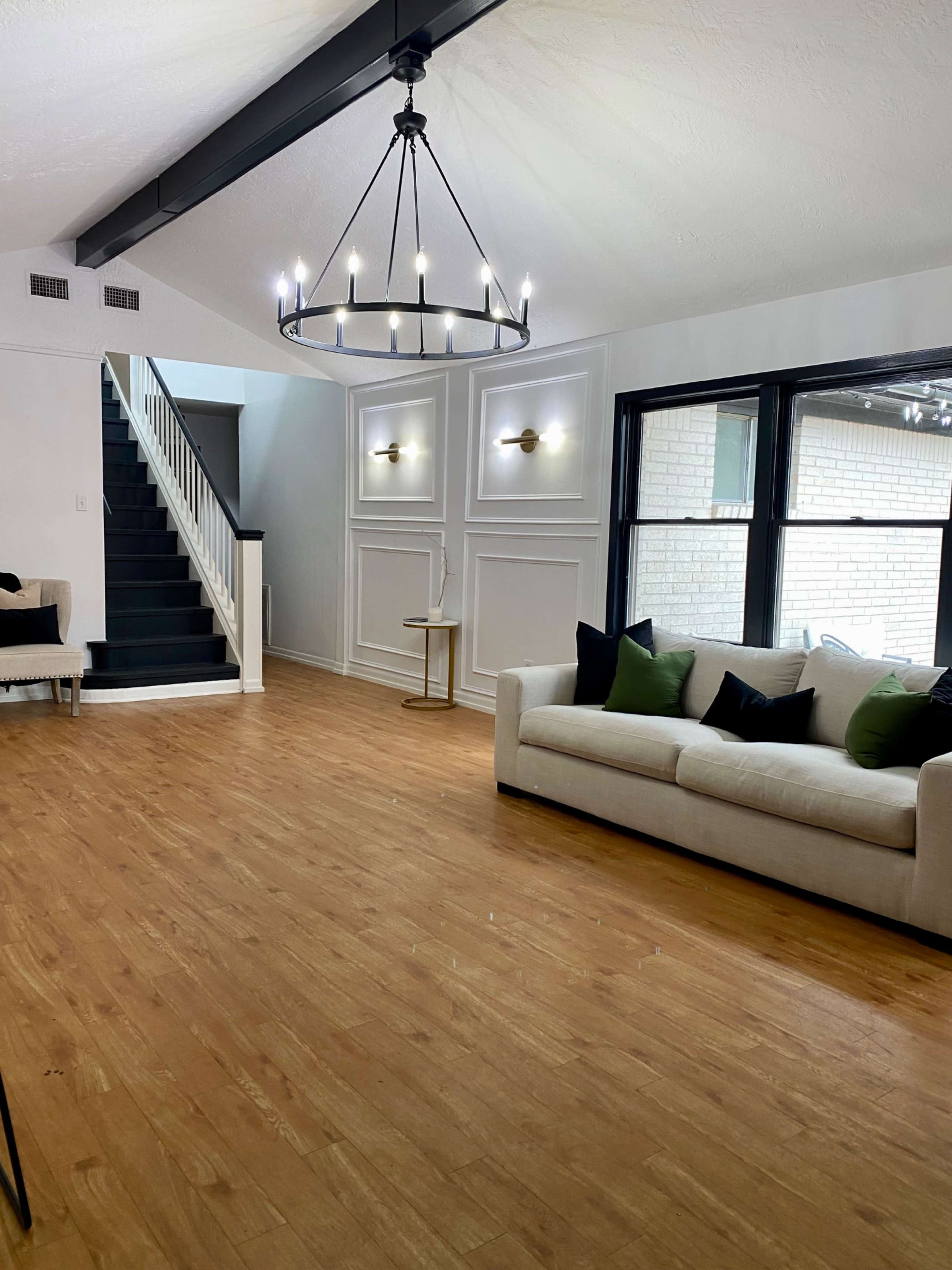 The living room features a chandelier above a beige sofa with green pillows, hardwood flooring, and a staircase leading to a second level.