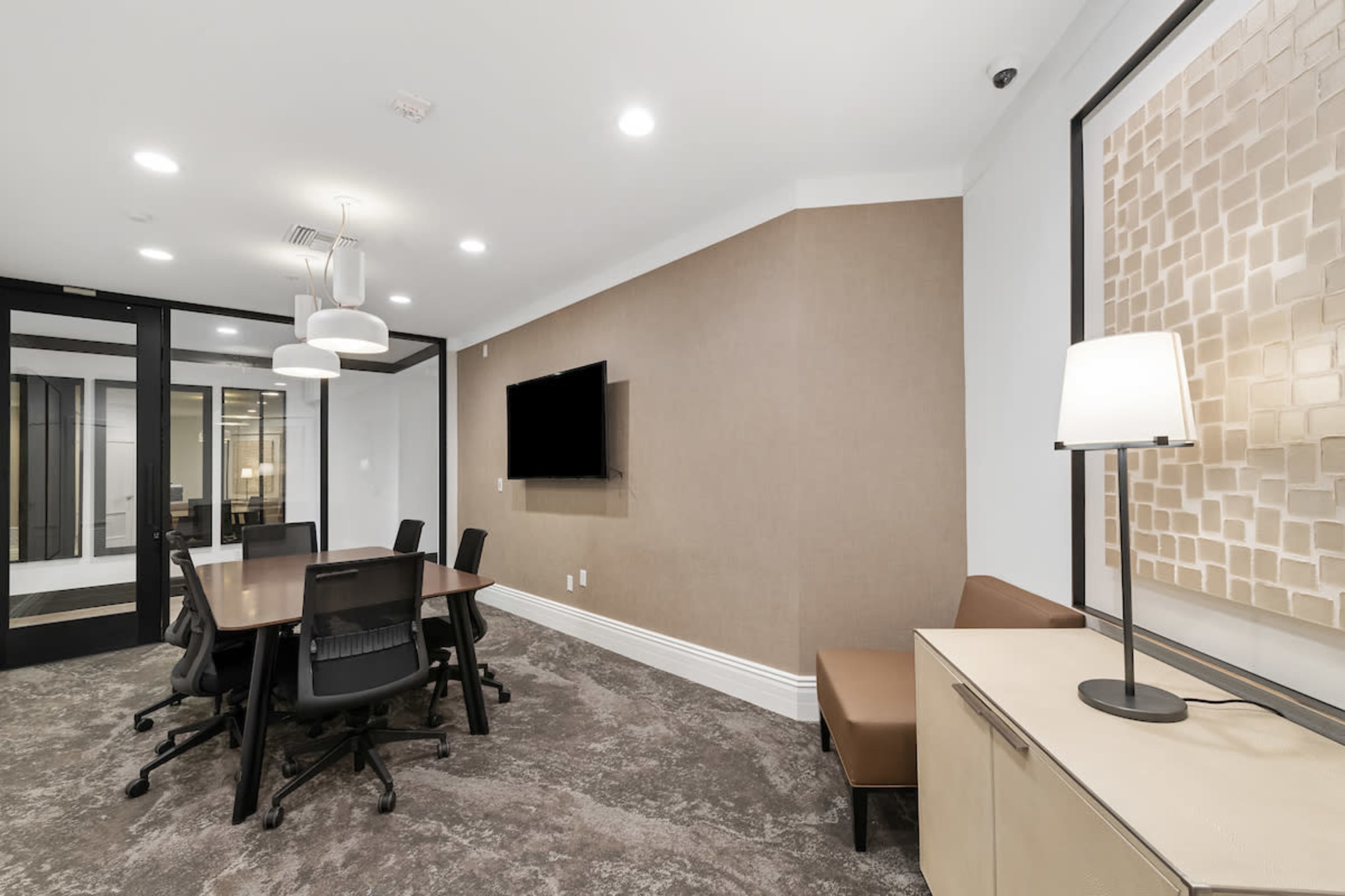 A conference room features a large table surrounded by chairs, a mounted television, and a side seating area with a lamp.