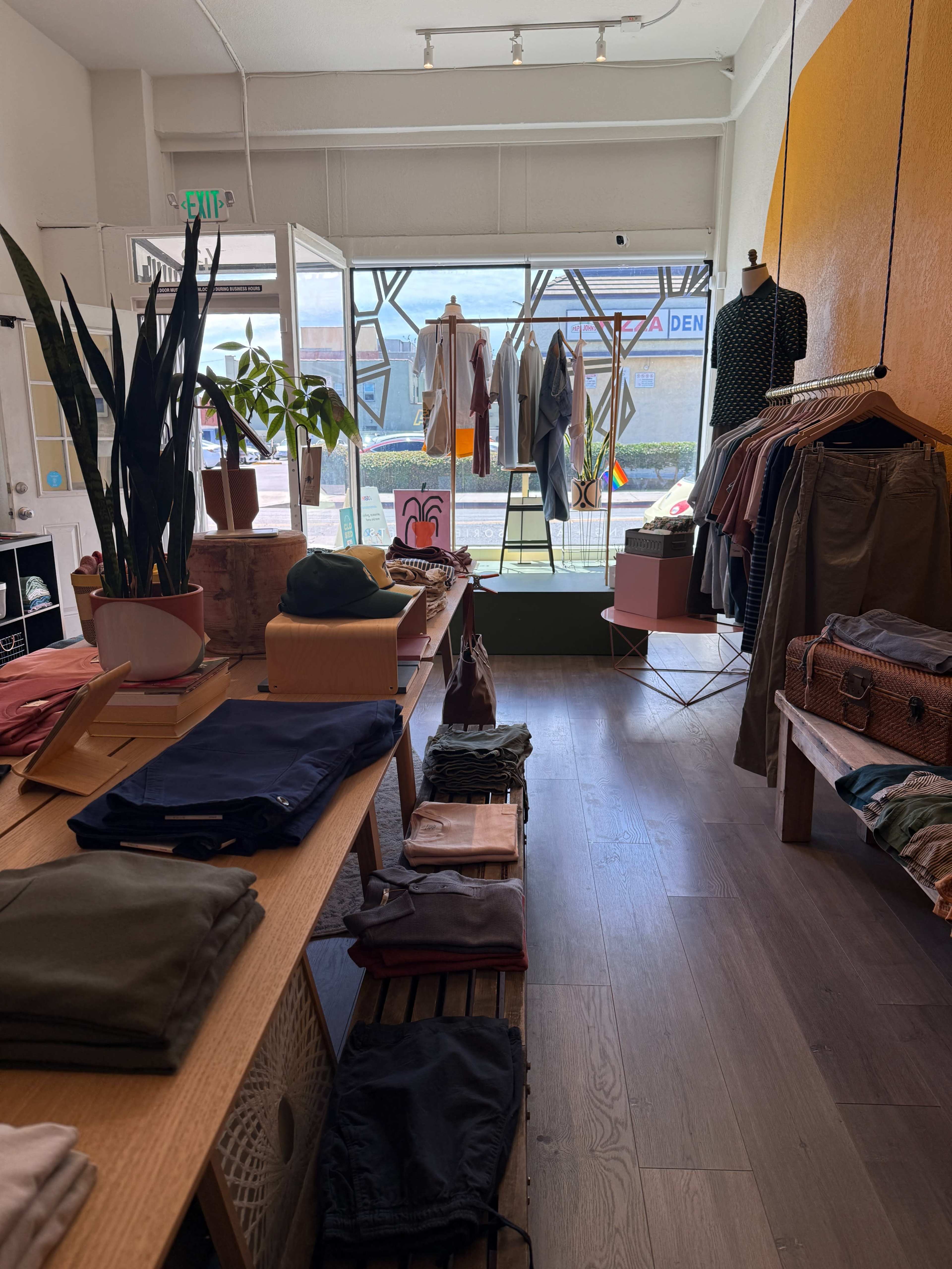 The image shows an interior view of a clothing store, featuring neatly arranged apparel on racks and tables, with a large window displaying additional clothing items.