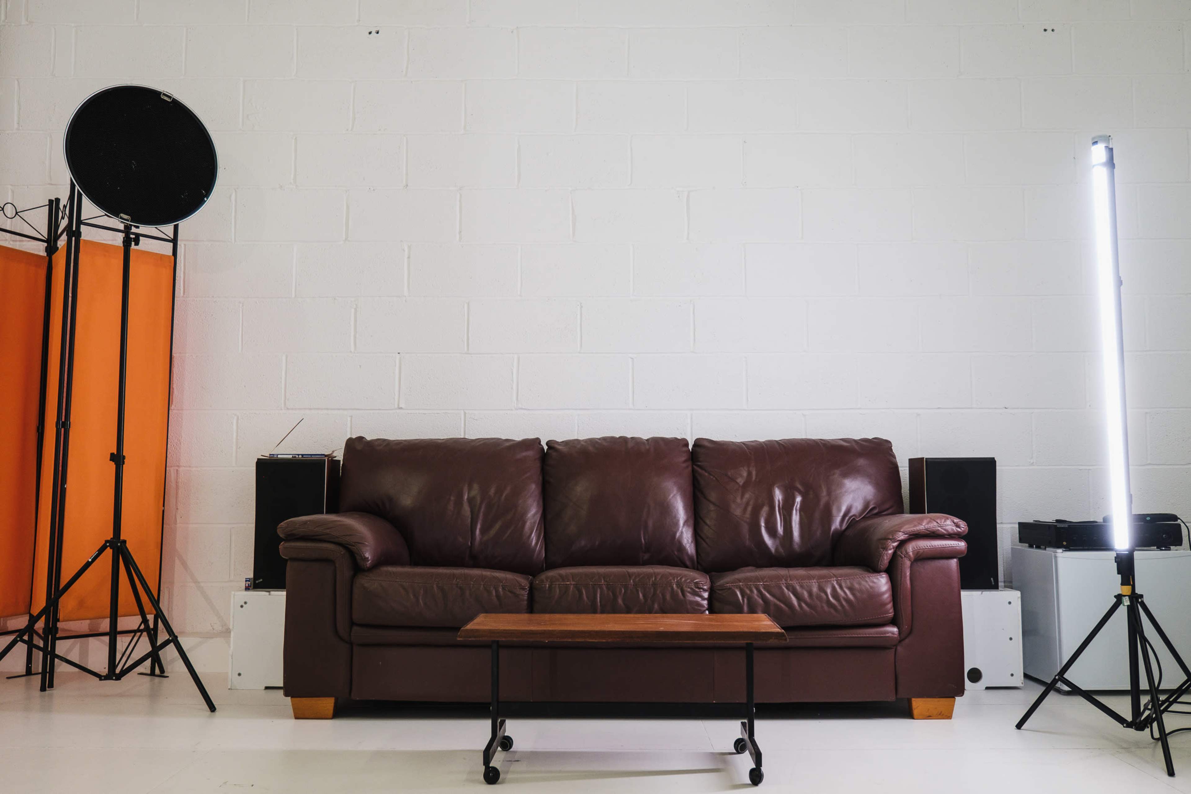 A brown leather couch centered against a white wall, with a small wooden coffee table in front and lighting equipment on either side.