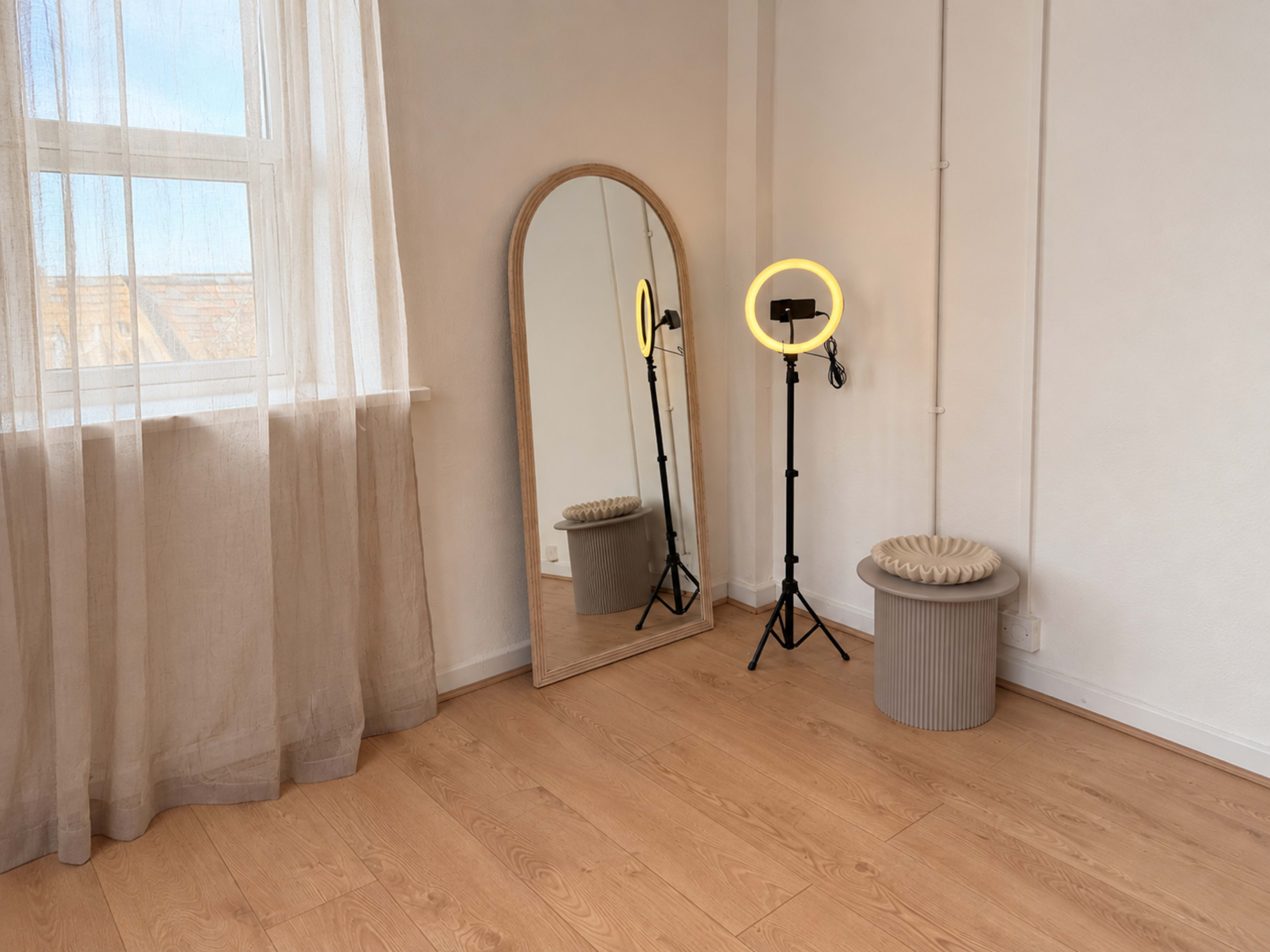 The image shows a minimalist room featuring a large mirror, a ring light on a tripod, a small round stool, and sheer curtains by the window.