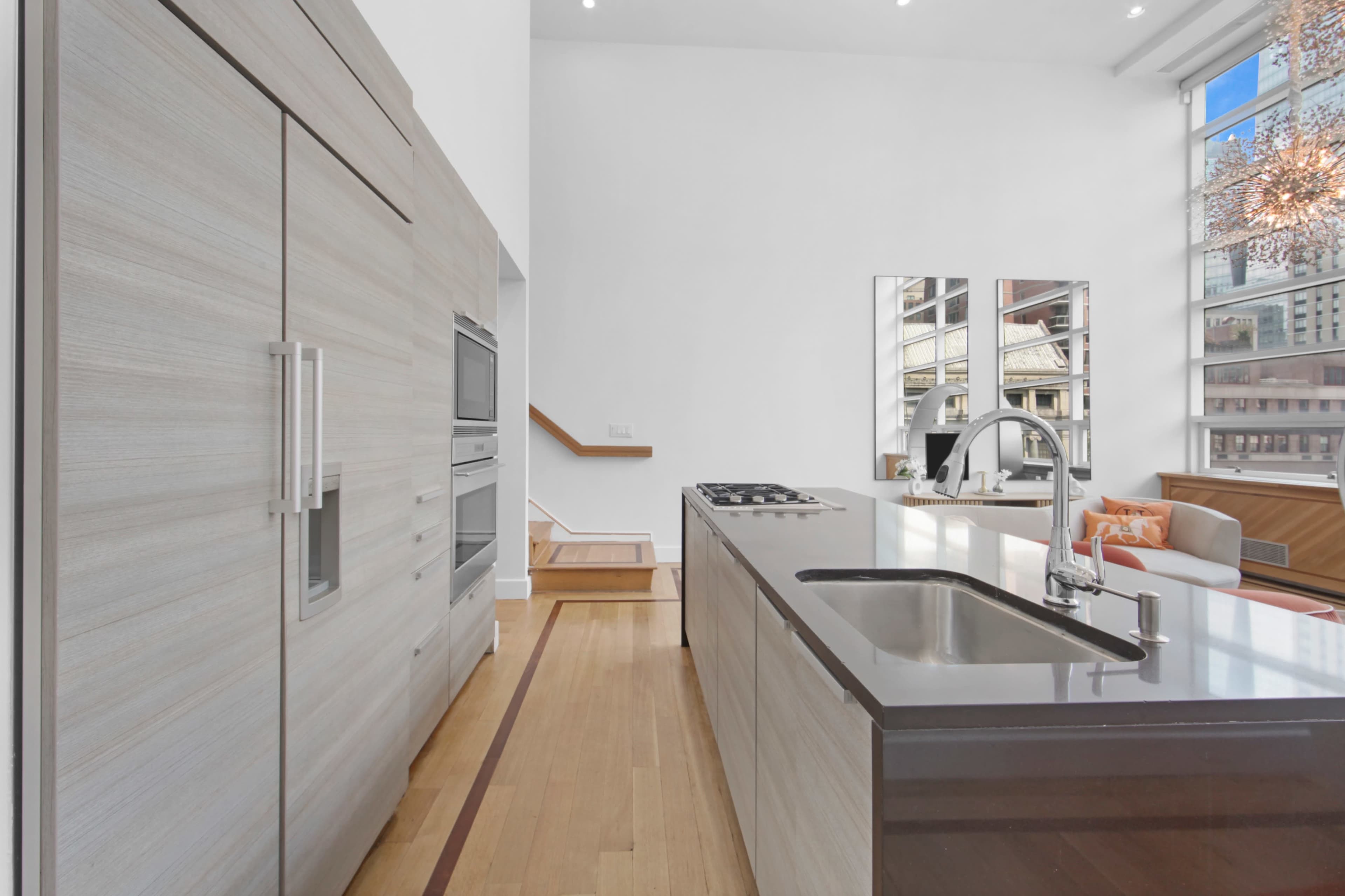 The image shows a modern kitchen with light-colored cabinetry, a central island, stainless steel appliances, and large windows allowing natural light to enter the space.